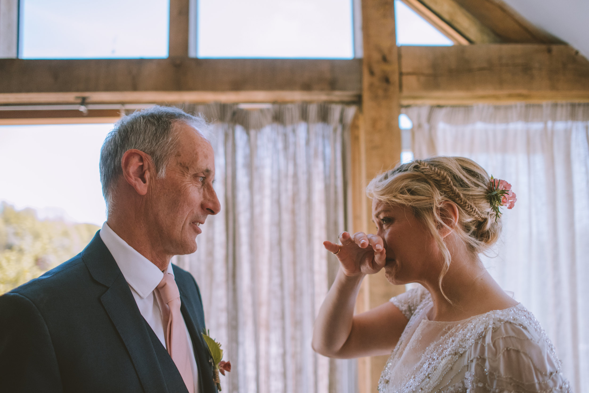 A bride with braided hair and a pink flower in her hair wiping tears away as she looks at a smiling groom dressed in a dark suit and white shirt inside a wooden venue with large windows.
