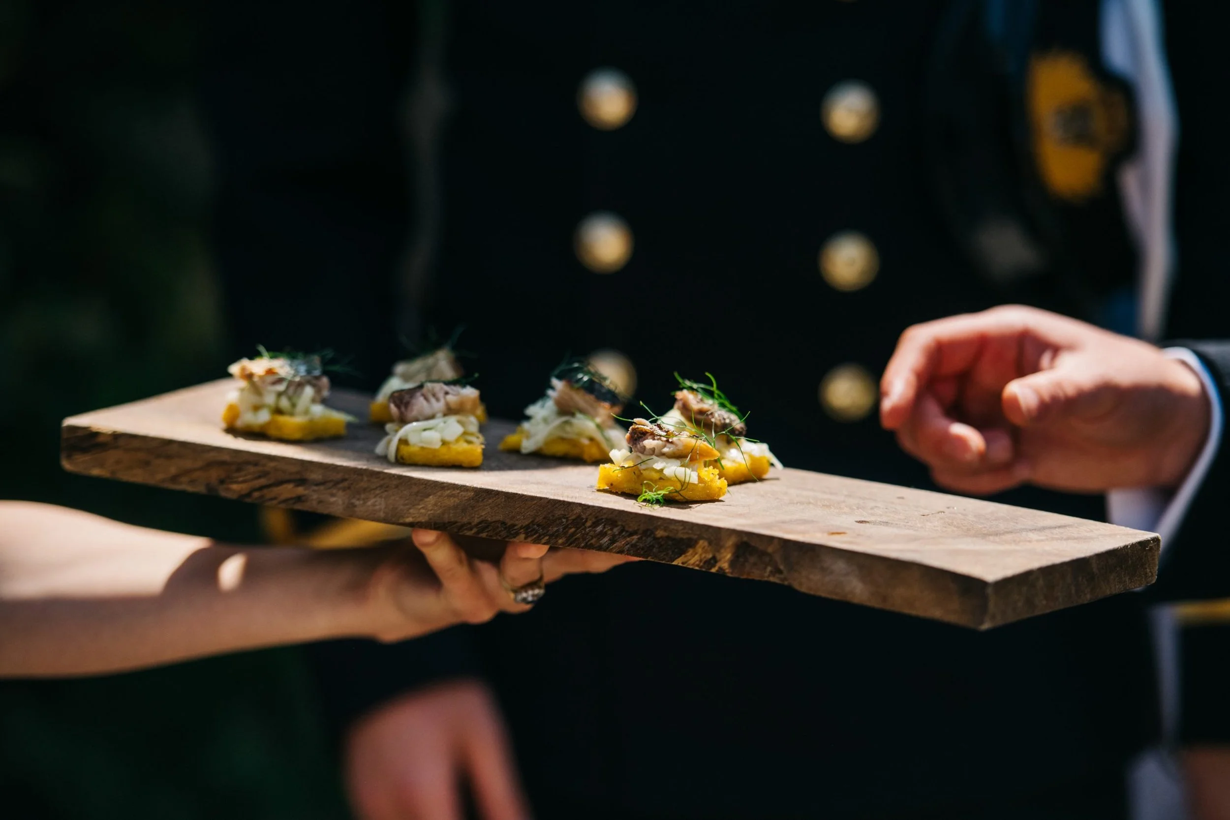 A person holding a wooden serving board with small gourmet appetizers garnished with herbs, during an outdoor event.