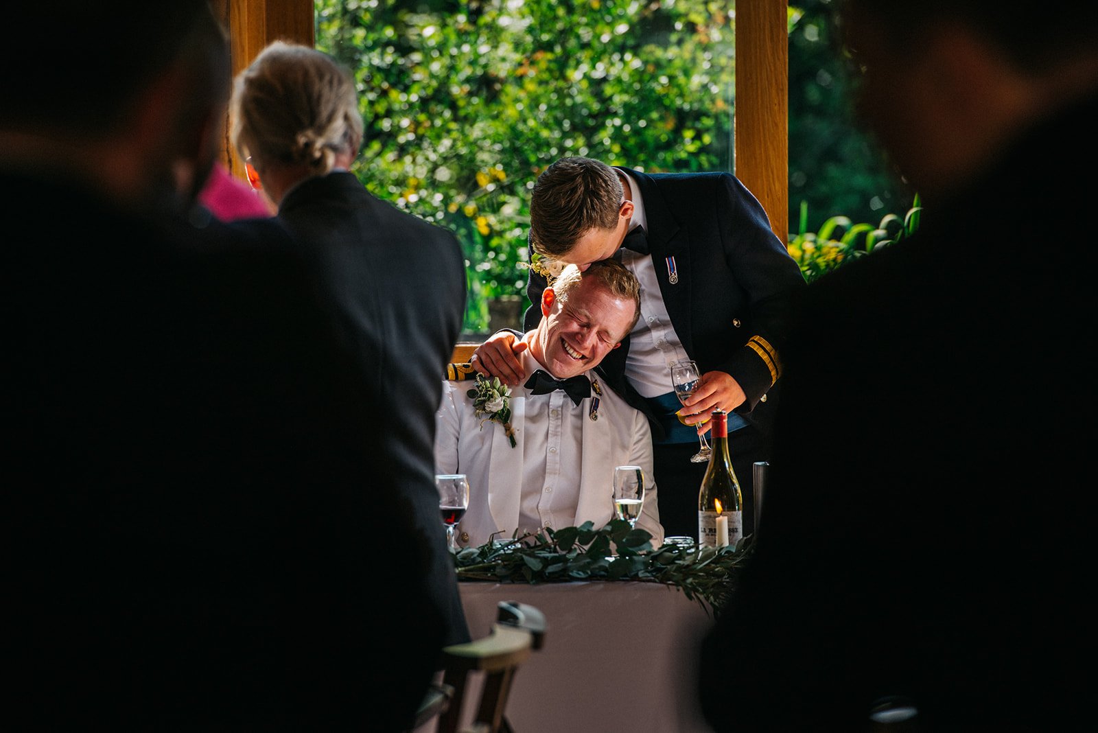 Two men in formal military attire sharing a joyful moment at a wedding reception, with one man seated and the other leaning over, both smiling and holding glasses of champagne. Guests are blurred in the foreground, and greenery is visible outside in 