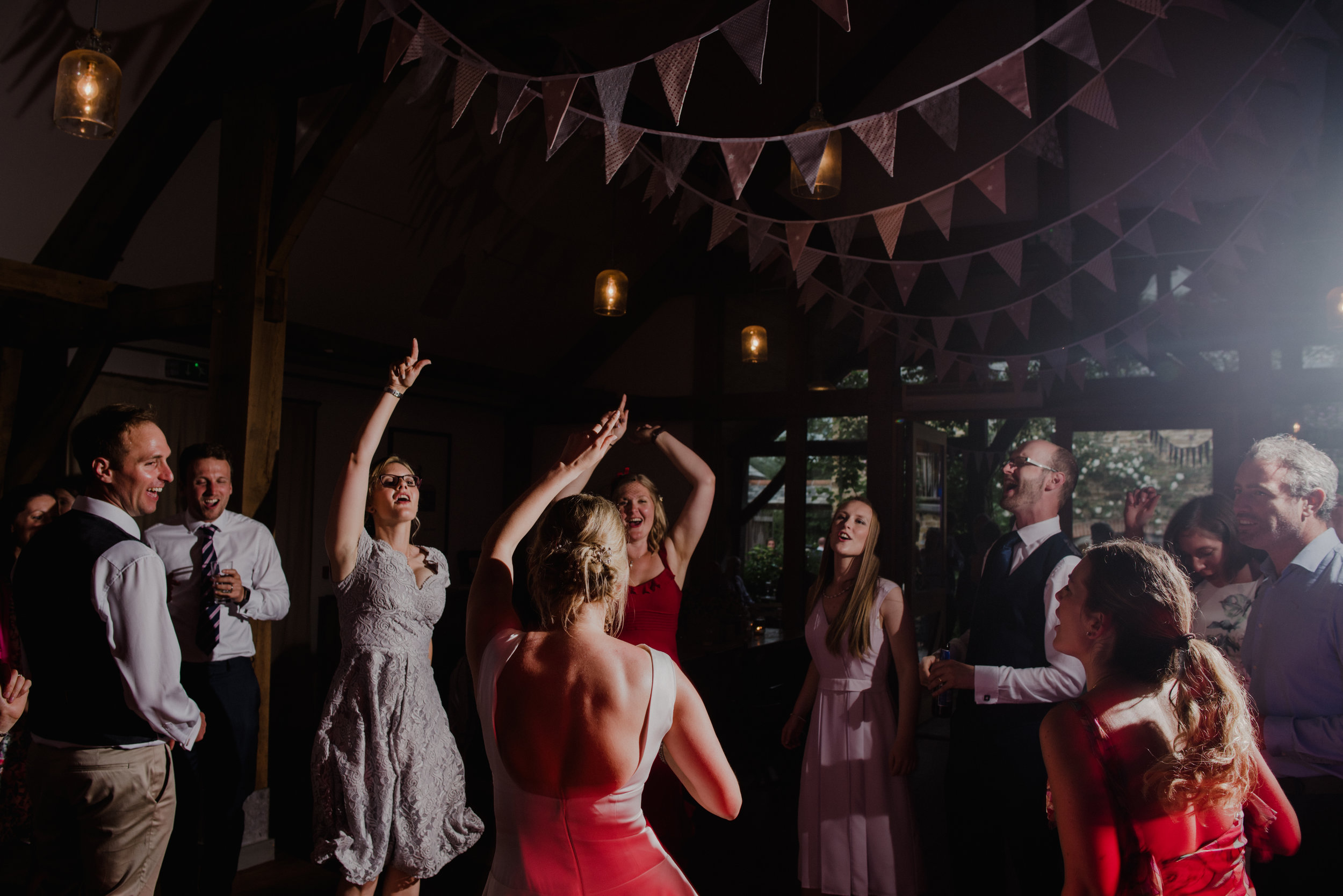 People dancing and celebrating at a party or wedding reception in a dimly lit venue decorated with bunting banners and hanging lights.
