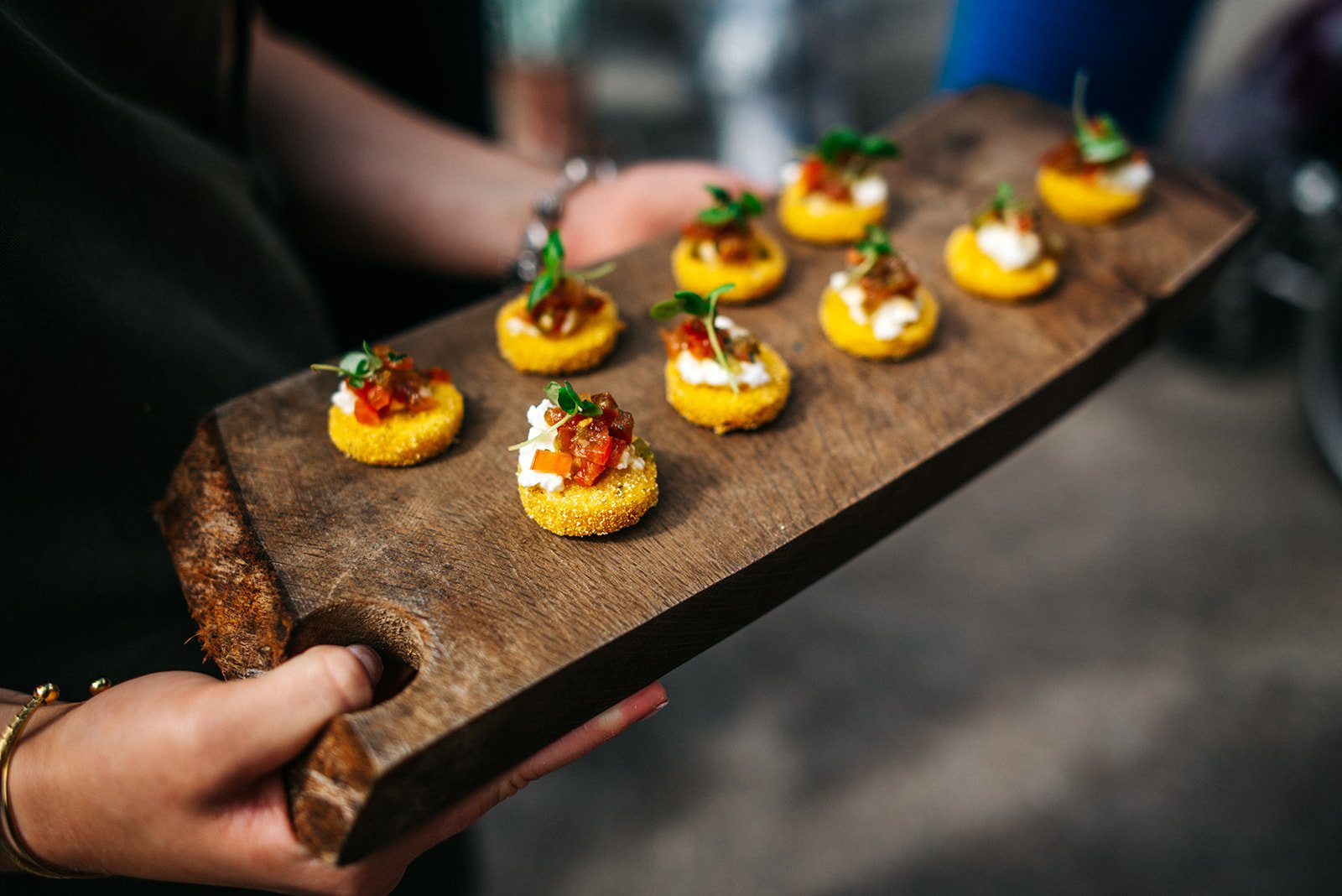 Person holding a wooden serving board with small, round, yellow-topped appetizers topped with garnishes.
