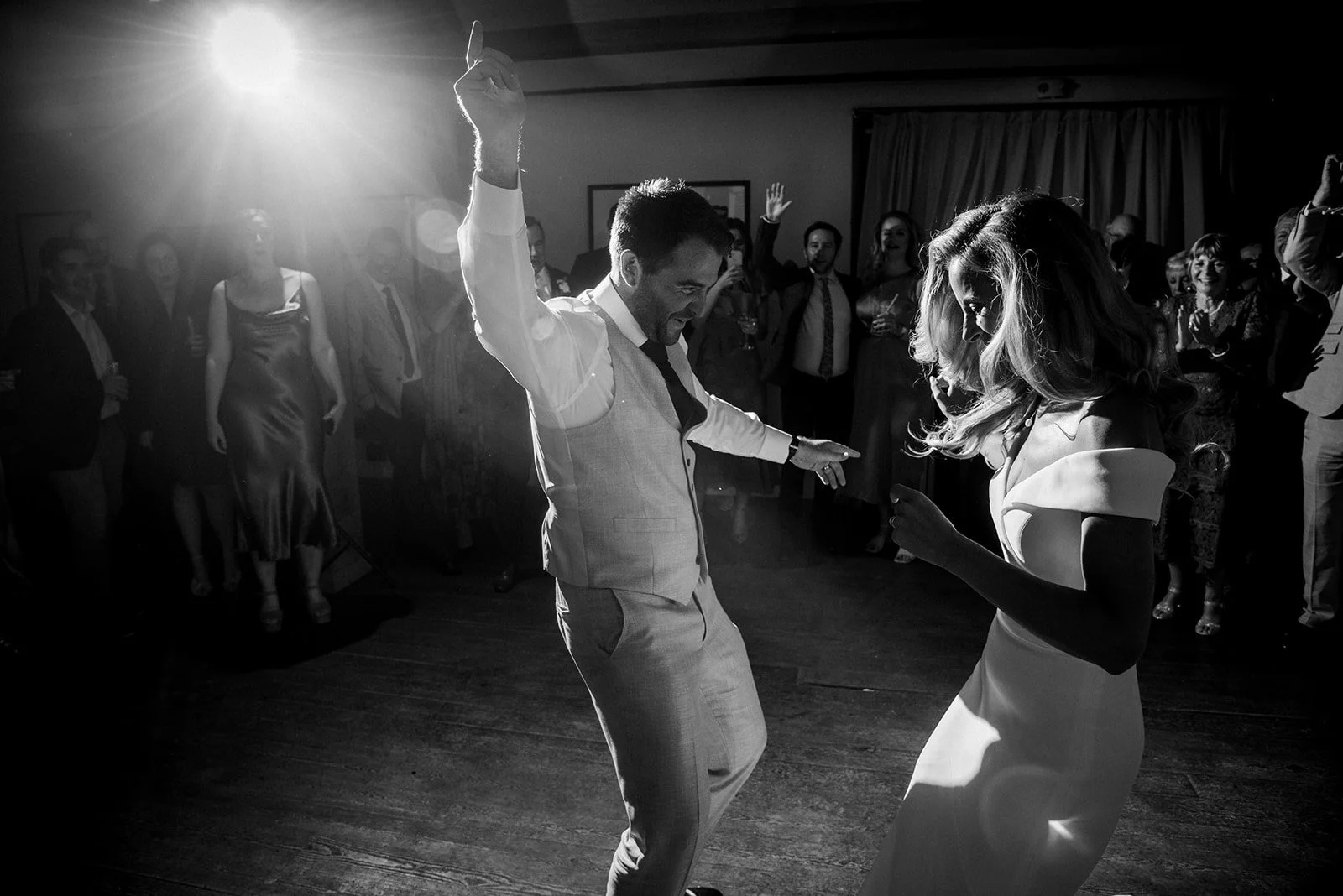 A black and white photo of a man and woman dancing at a wedding reception, surrounded by guests.