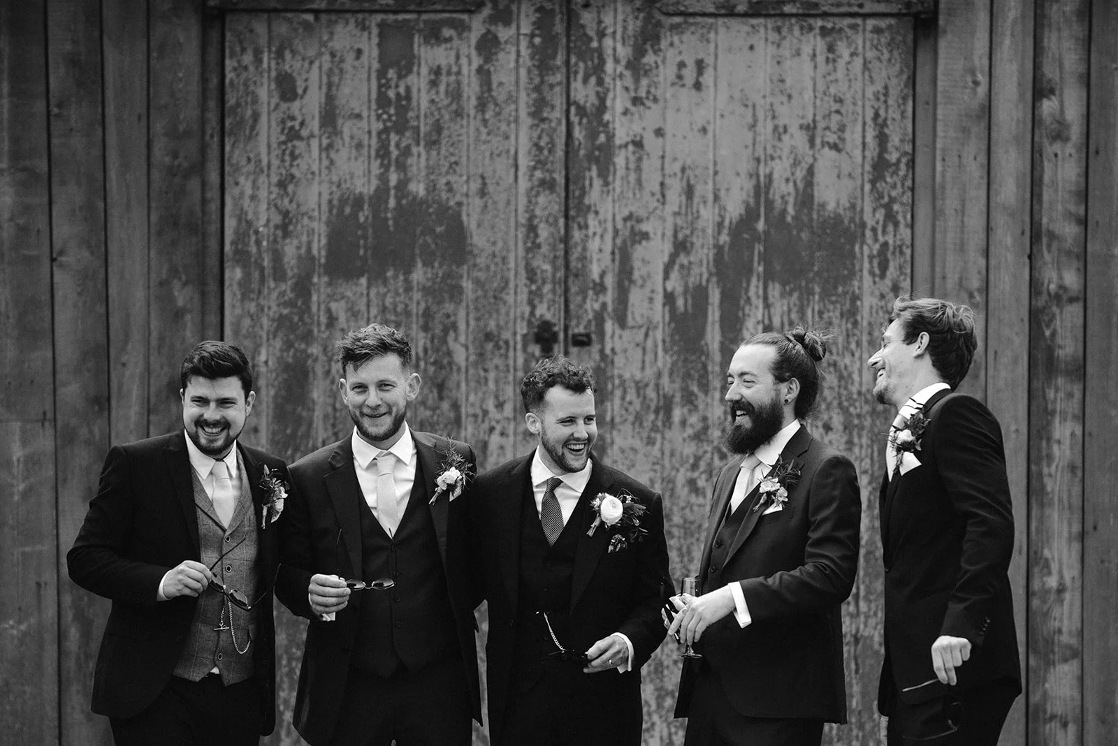 Five men in tuxedos laughing and smiling together at a wedding reception or event, with a rustic wooden background.