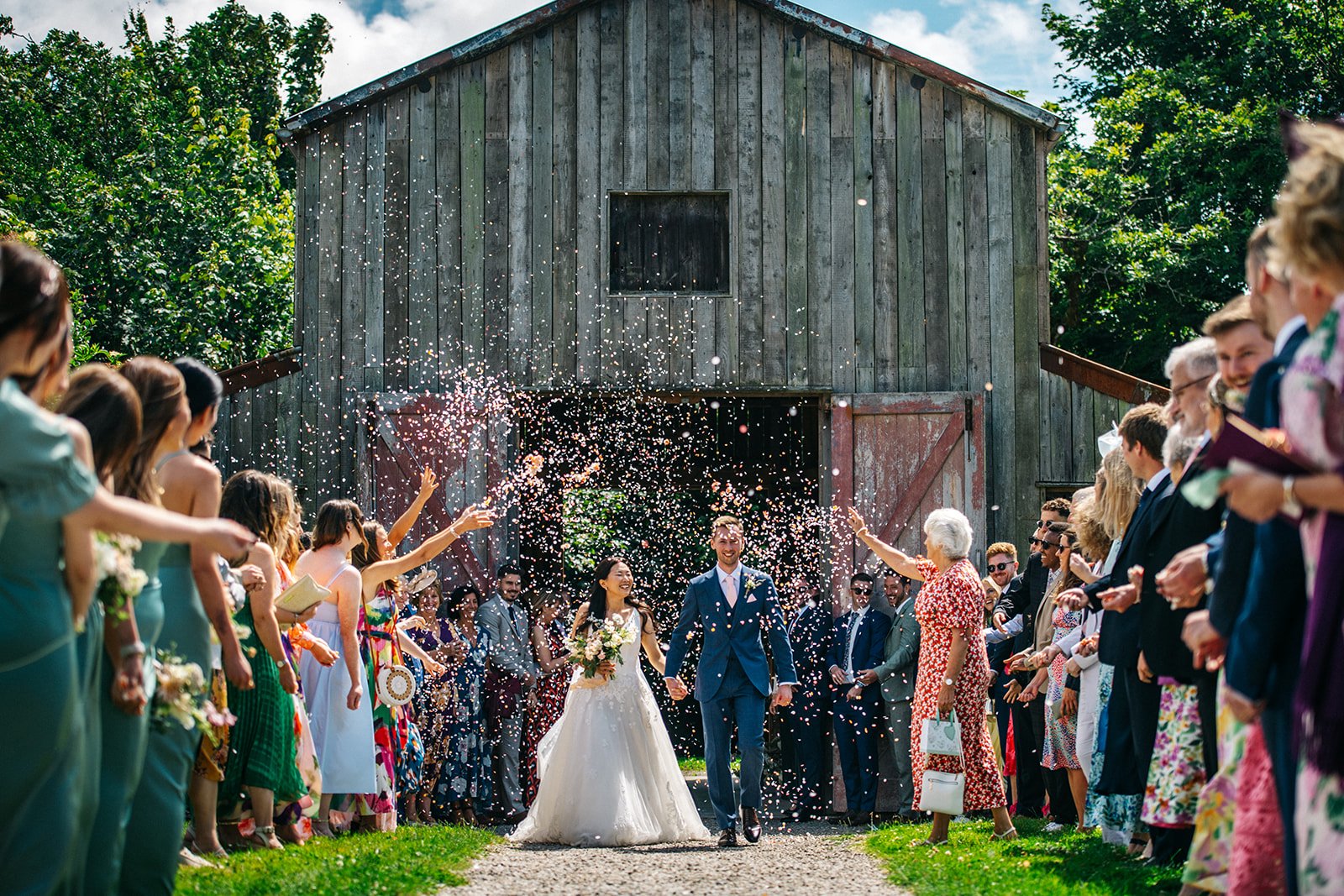 A wedding procession with a bride and groom walking hand-in-hand through a shower of confetti in front of a rustic barn, surrounded by guests celebrating outdoors.