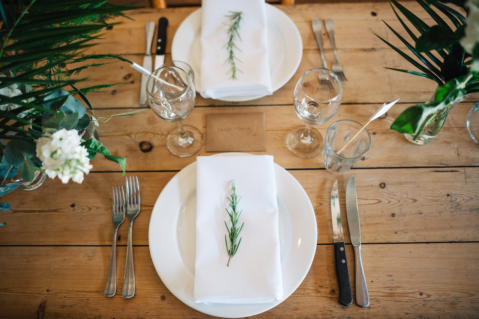 A rustic wooden dining table set for two with white plates, folded white napkins, silverware, empty wine glasses, and small vases with greenery and white flowers, decorated with sprigs of greenery.