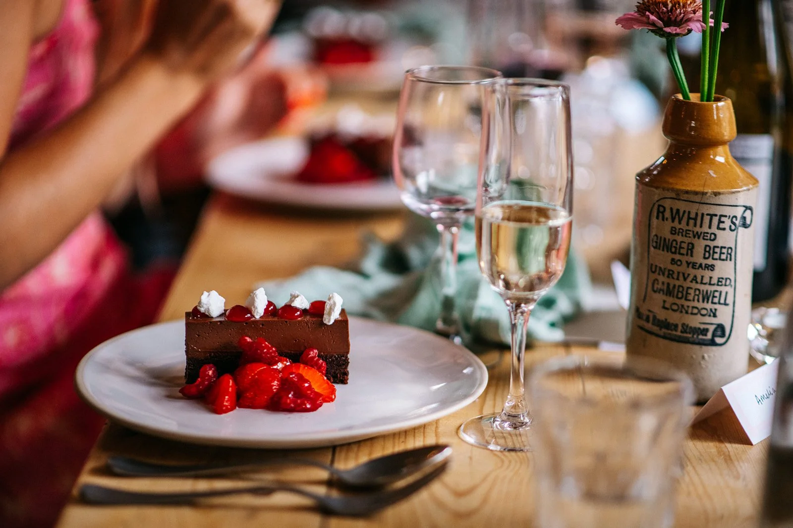 Chocolate cake with berries and whipped cream on a white plate, two wine glasses, and a decorative bottle with flowers on a wooden table at a celebration or gathering.