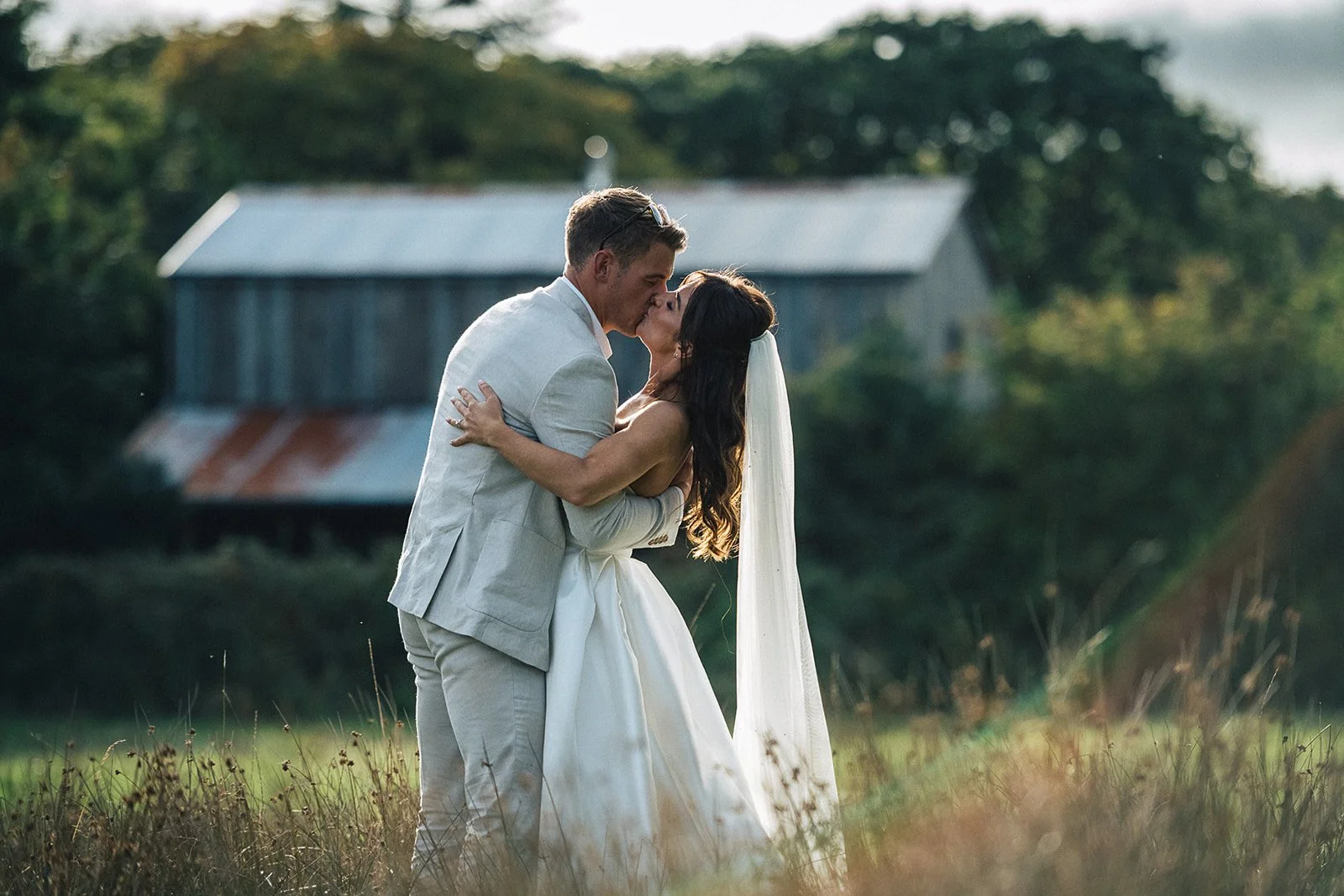 A couple in wedding attire sharing a kiss outdoors in a field with a rustic barn in the background.