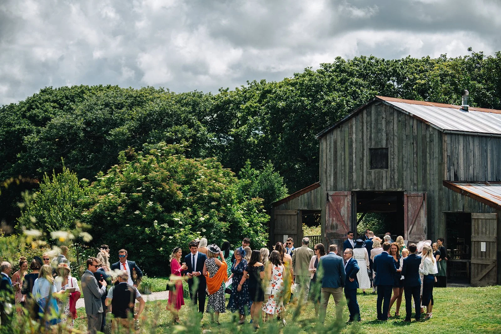 A group of people dressed in formal and semi-formal attire gathered outside a rustic barn on a cloudy day. The scene appears to be a wedding or special event.