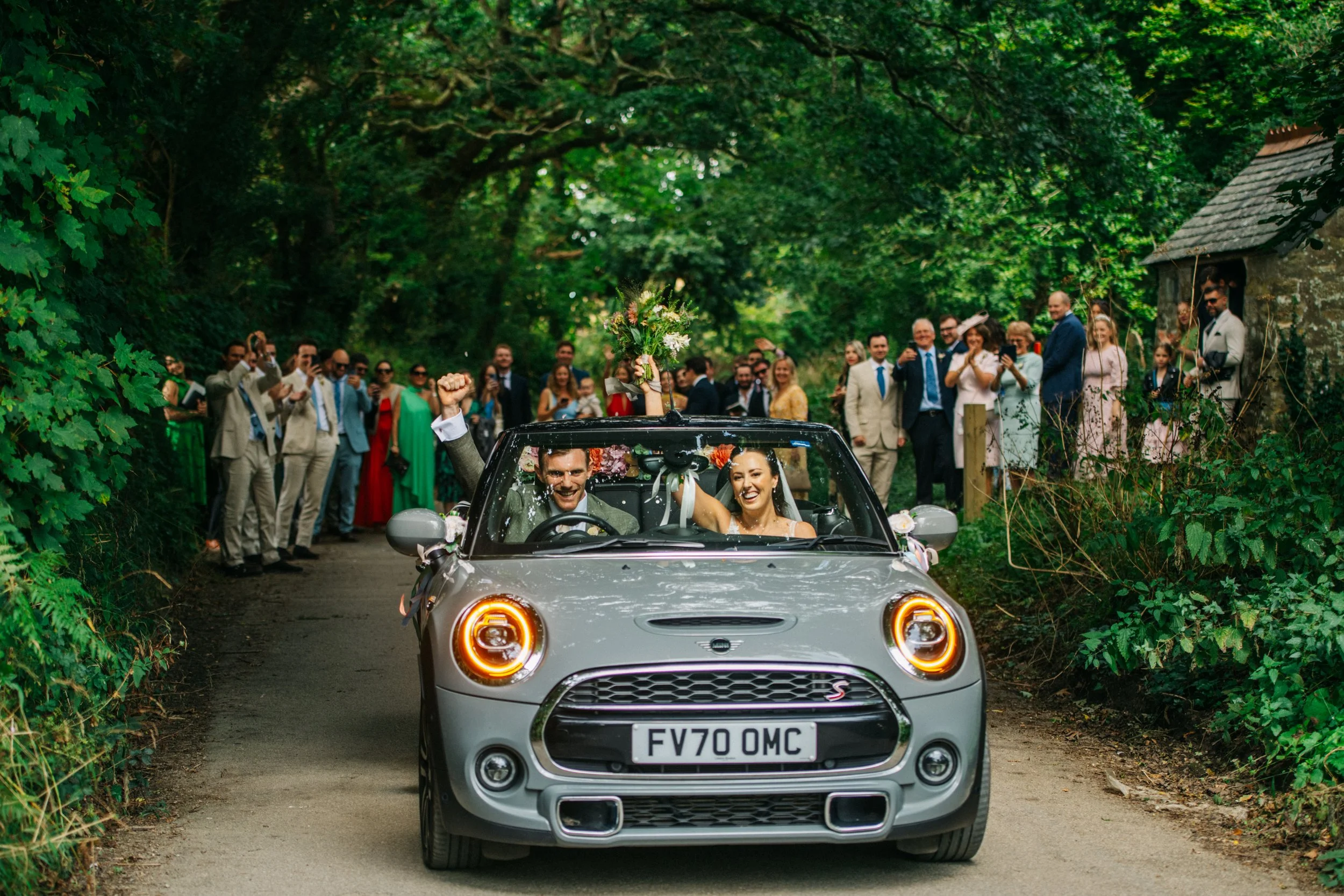 A newly married couple driving in a silver Mini Cooper convertible through a wooded area, with a crowd of friends and family celebrating and taking photos in the background.