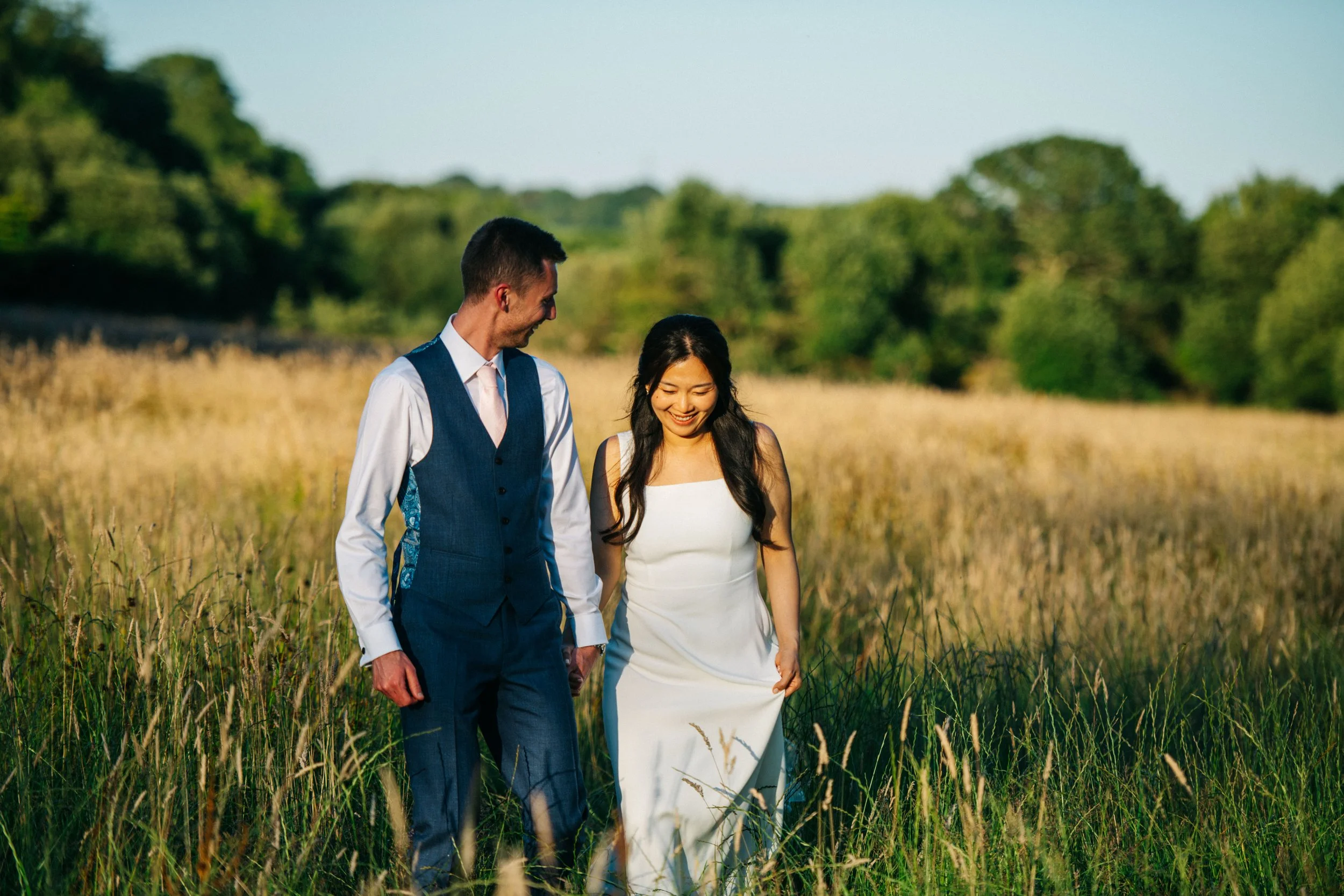 A newlywed couple walking through a grassy field on a sunny day, smiling and enjoying each other's company.