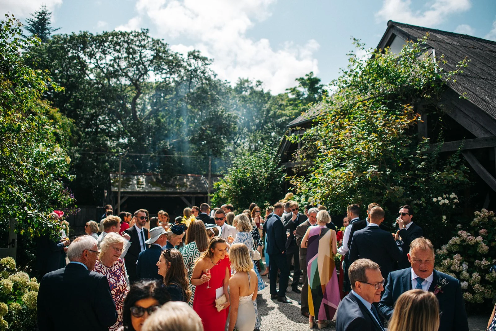 A group of people at an outdoor gathering or party, dressed in formal and semi-formal attire, amidst greenery and rustic buildings.