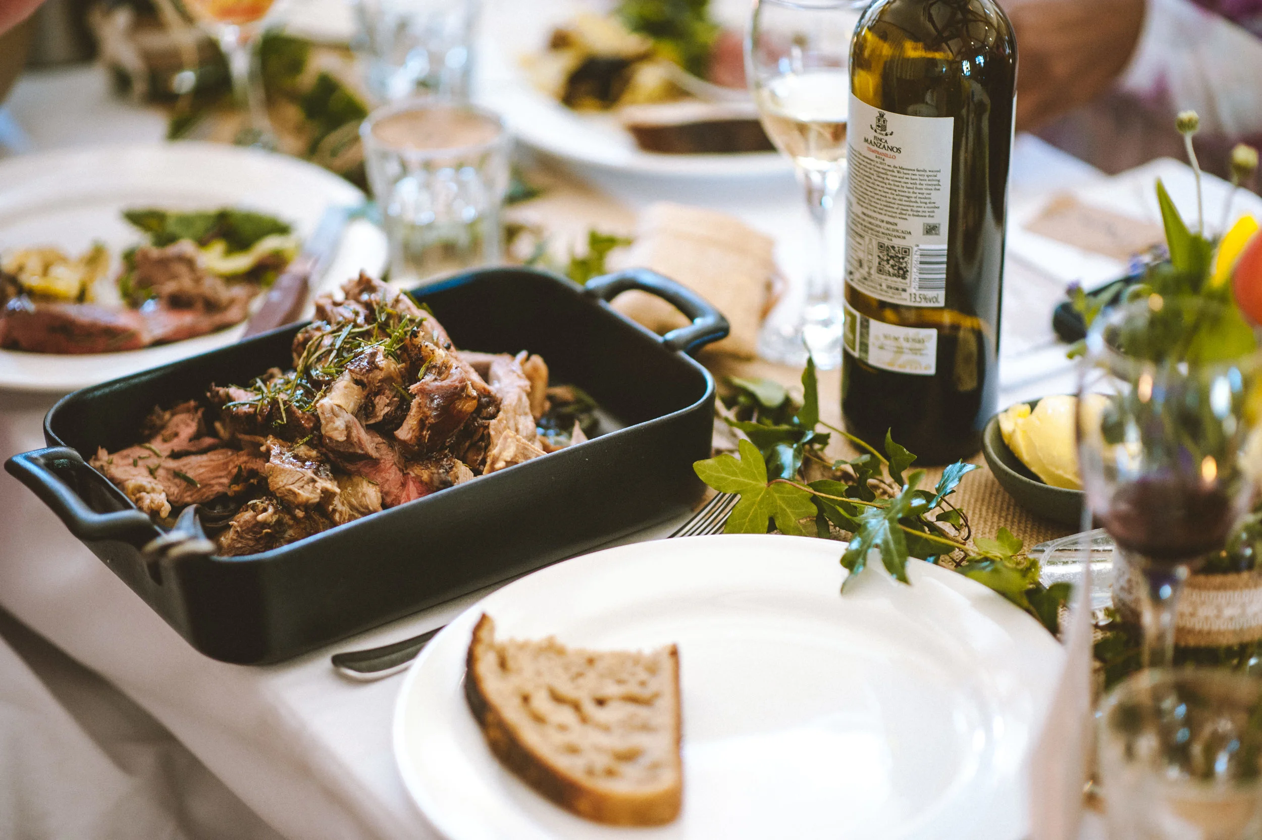 A table set for a meal with a dish of roasted meat garnished with herbs, a bottle of wine, wine glasses, a partially eaten bread, and various plates and glasses, decorated with greenery and flowers.