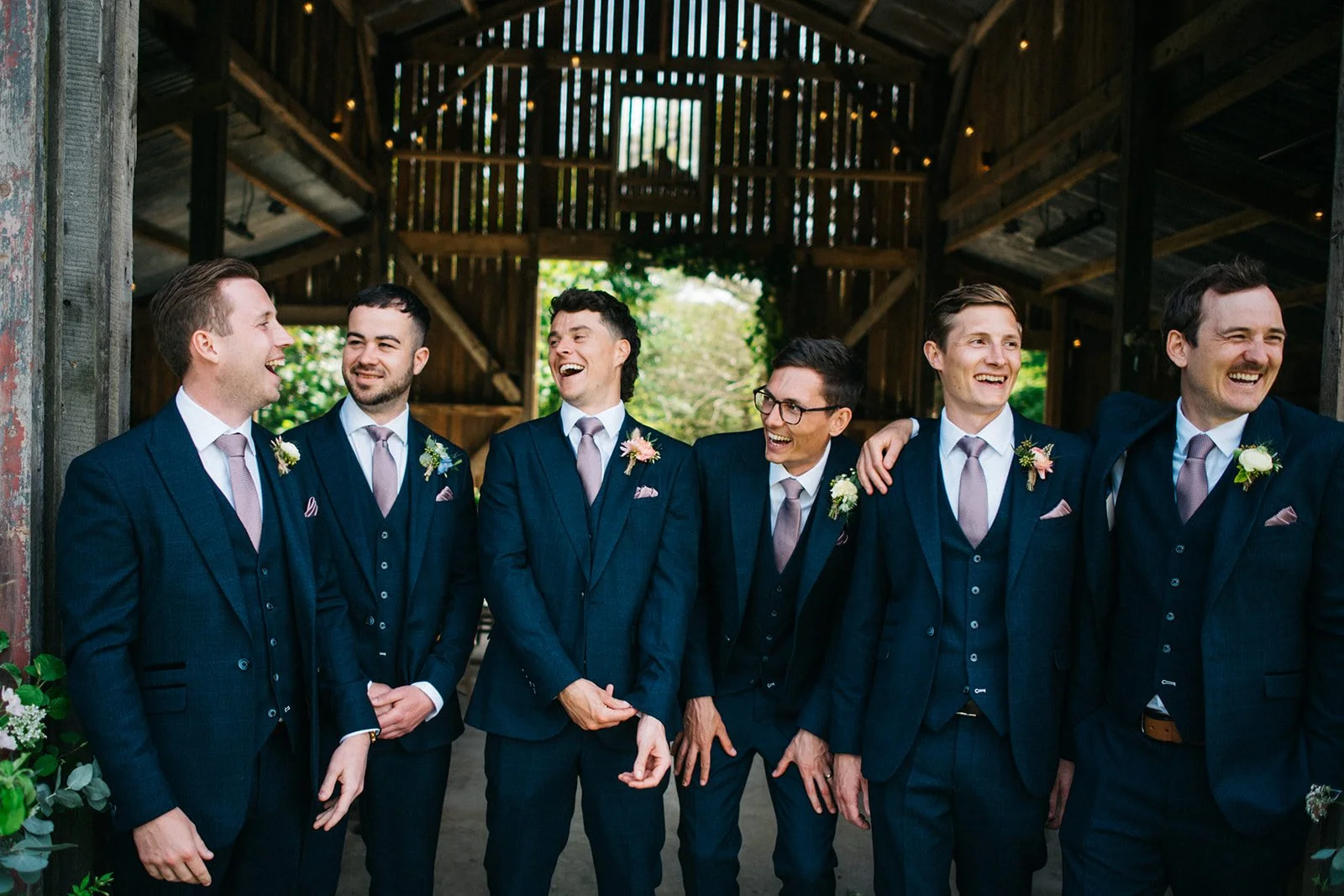 A group of six men in navy suits with pink ties and boutonnieres, smiling and laughing together inside a rustic barn with wood walls and open windows, celebrating a wedding.