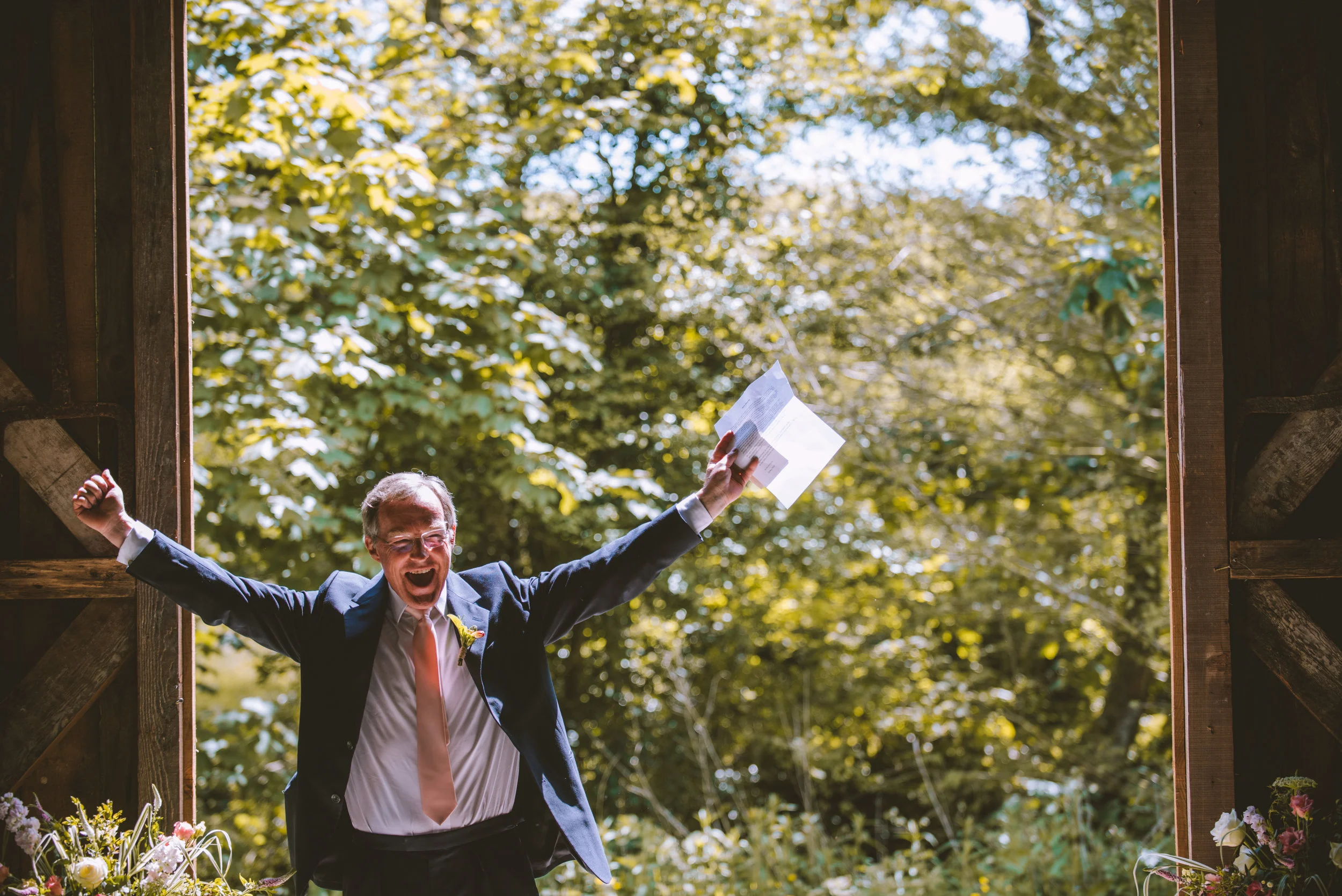 A man in a suit celebrating with arms outstretched, holding a piece of paper, outside with a background of leafy trees.