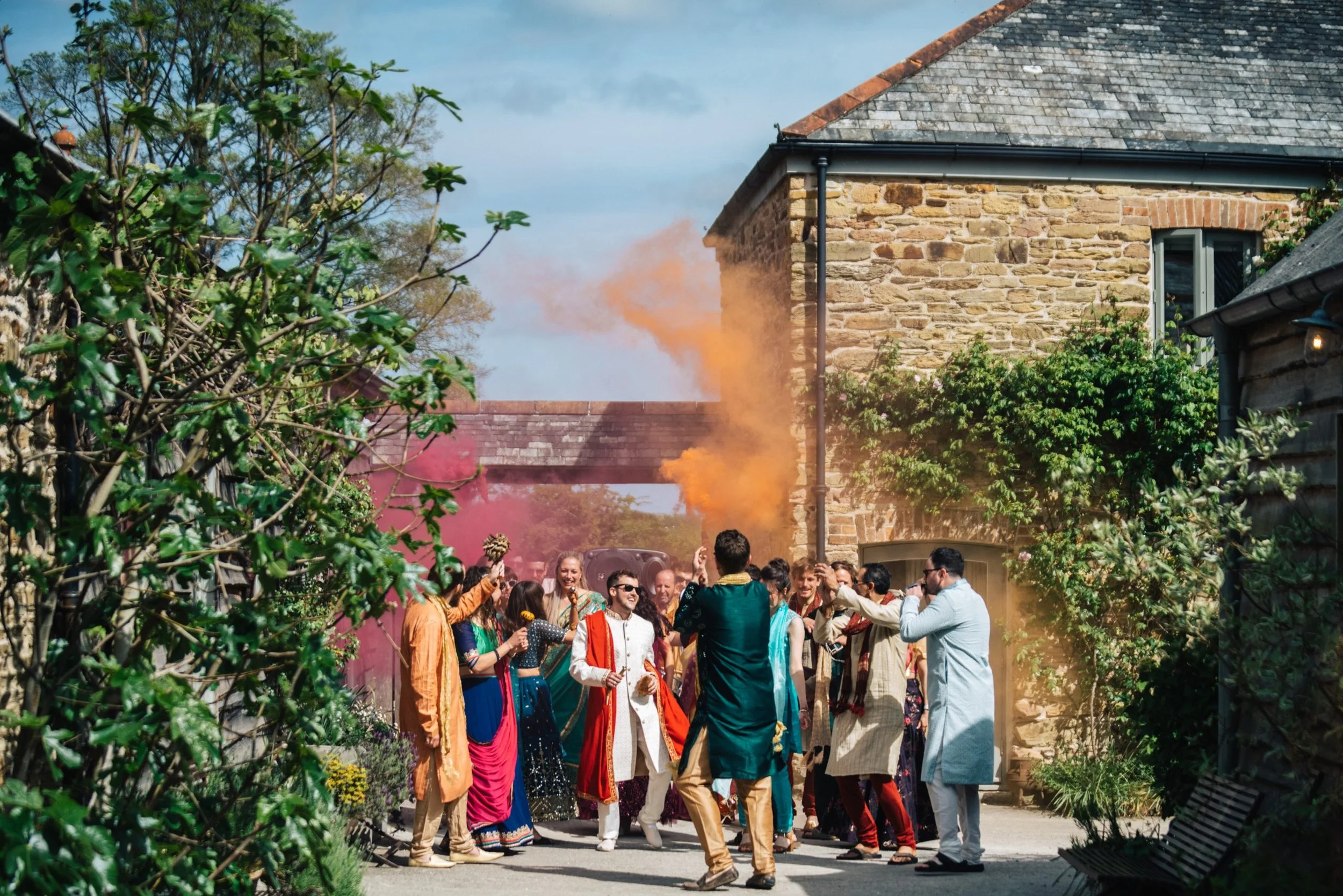 A group of people dressed in traditional Indian attire celebrating outdoors with colorful smoke, smiling and dancing near stone buildings and greenery.