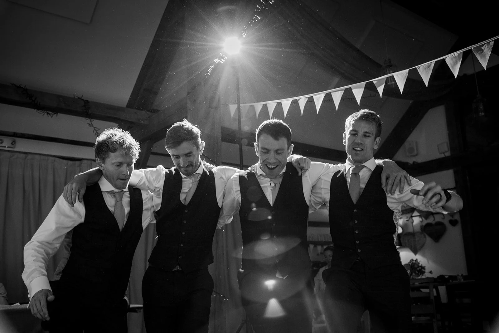 Four men in formal attire with arms around each other's shoulders, dancing together at a celebration or party in a decorated indoor venue.