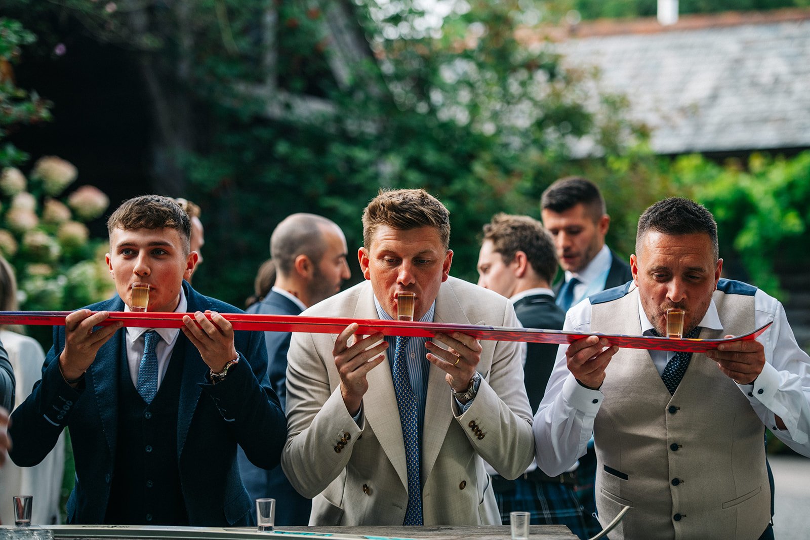 Three men in formal attire appear to be drinking champagne from glasses balanced on a red ribbon at an outdoor event.