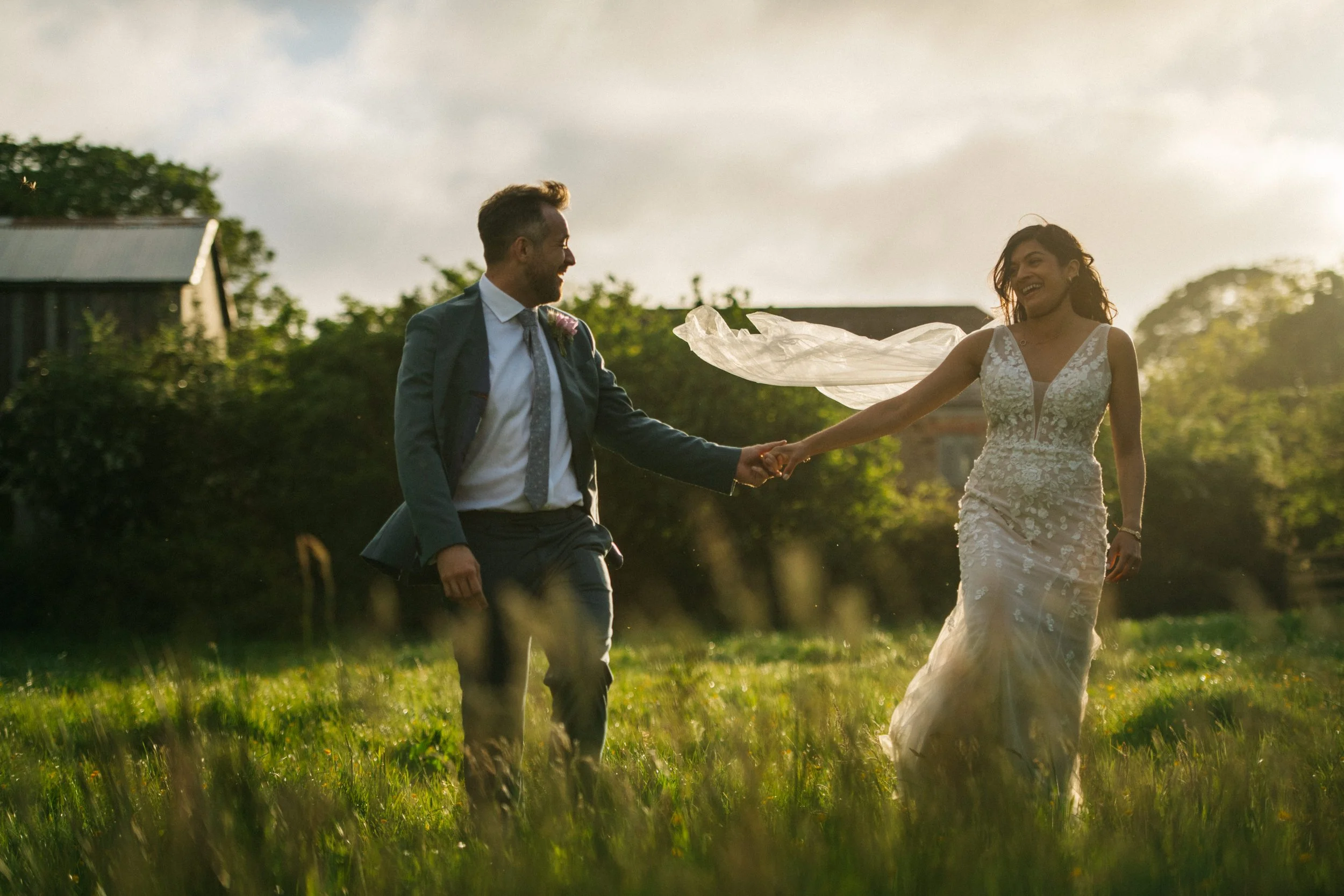 A bride and groom holding hands and dancing in a green field during sunset. The bride is wearing a white lace wedding dress, and the groom is wearing a suit. The bride's veil is flowing in the wind, and they are smiling at each other.