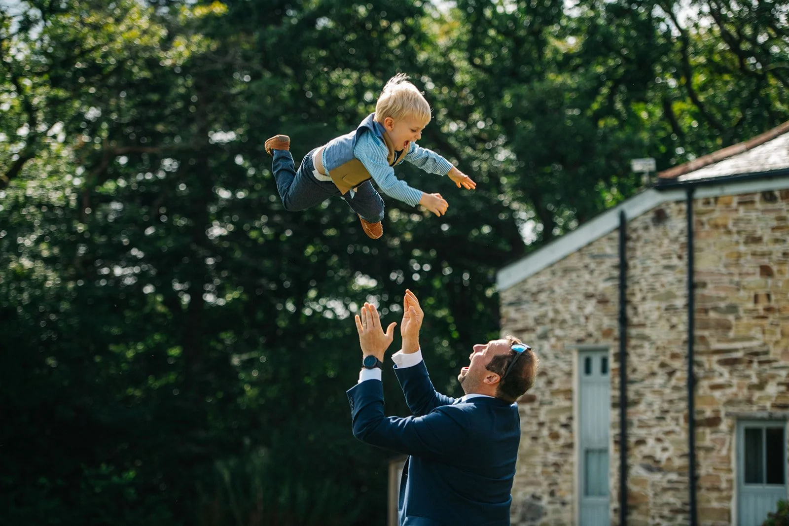 A man in a suit and sunglasses is tossing a boy in a blue outfit into the air outdoors with trees and a stone house in the background.