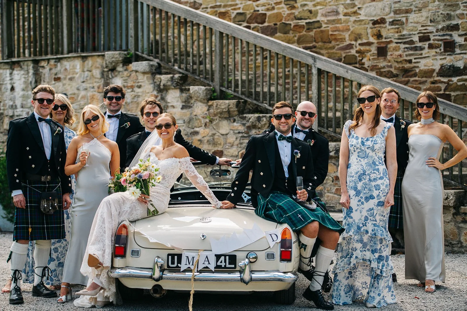 Group of wedding guests in formal attire posing around a vintage Mercedes car, with a bride in a lace wedding dress sitting on the trunk holding a bouquet, and others wearing kilts and sunglasses, in front of a stone wall and staircase.
