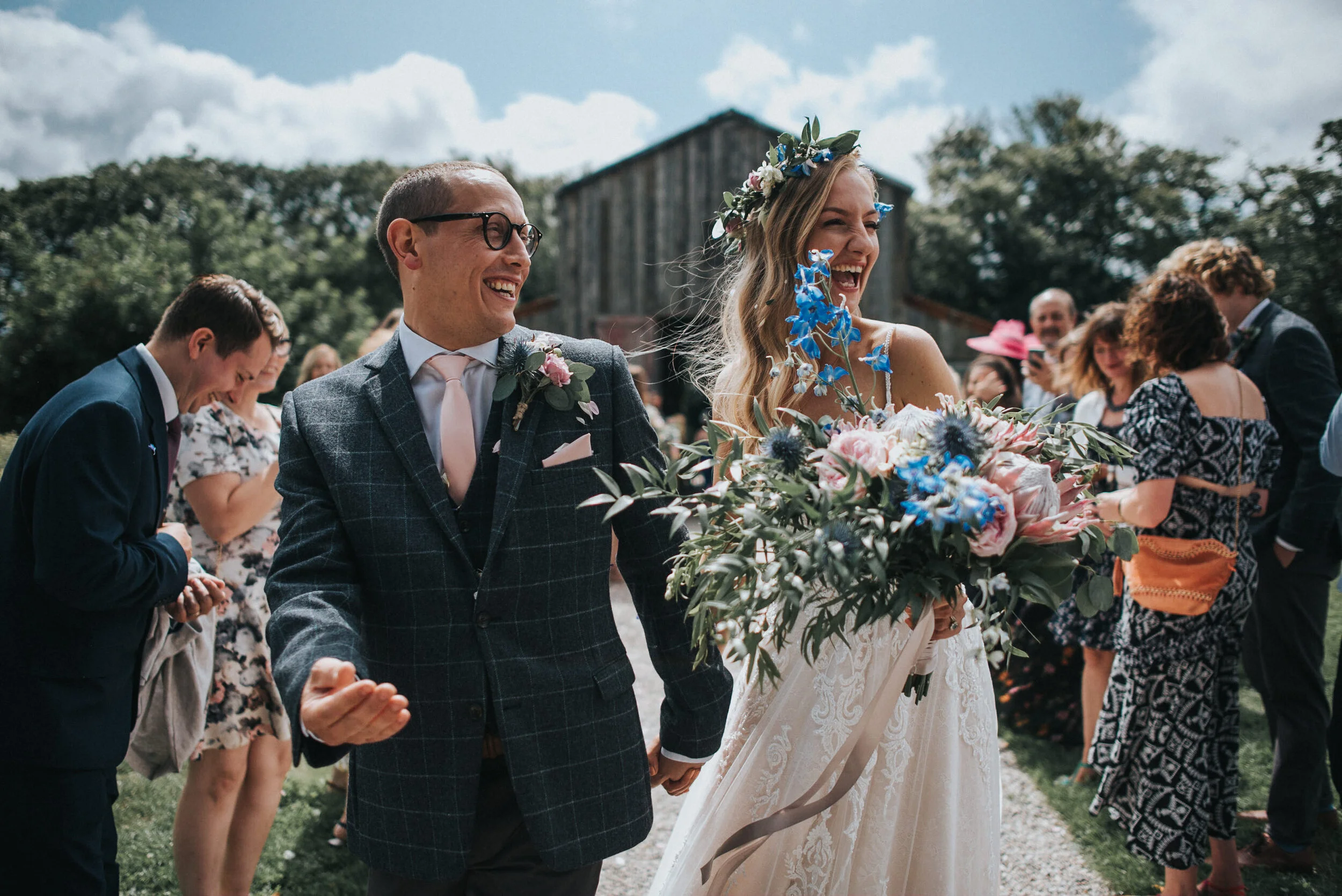 Happy bride in a white lace wedding dress and a floral crown holding a large bouquet of pink and blue flowers, walking outdoors with a groom in a plaid suit and glasses, surrounded by smiling guests.