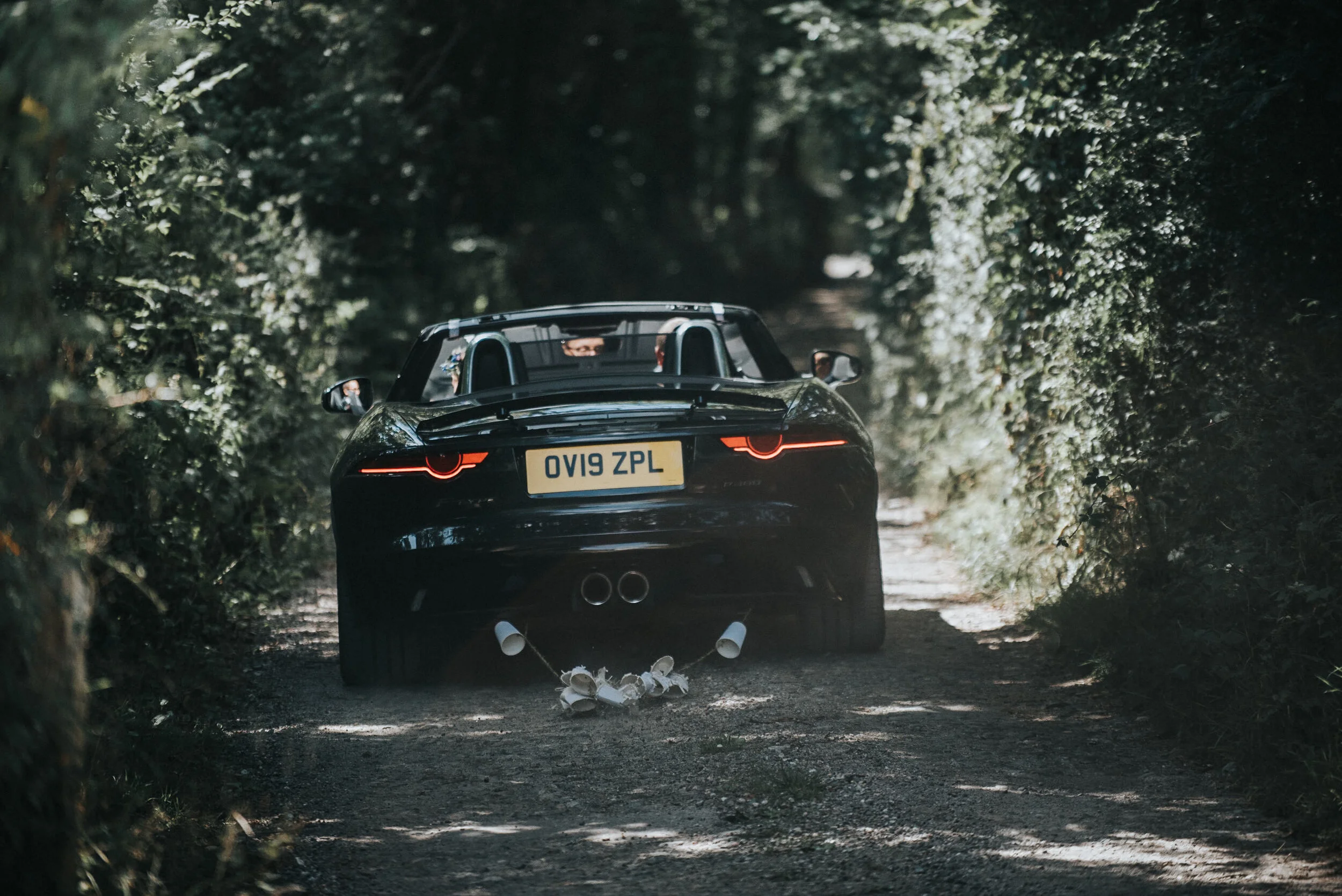A black convertible sports car driving on a gravel woodland path with disposable coffee cups hanging from the rear bumper.