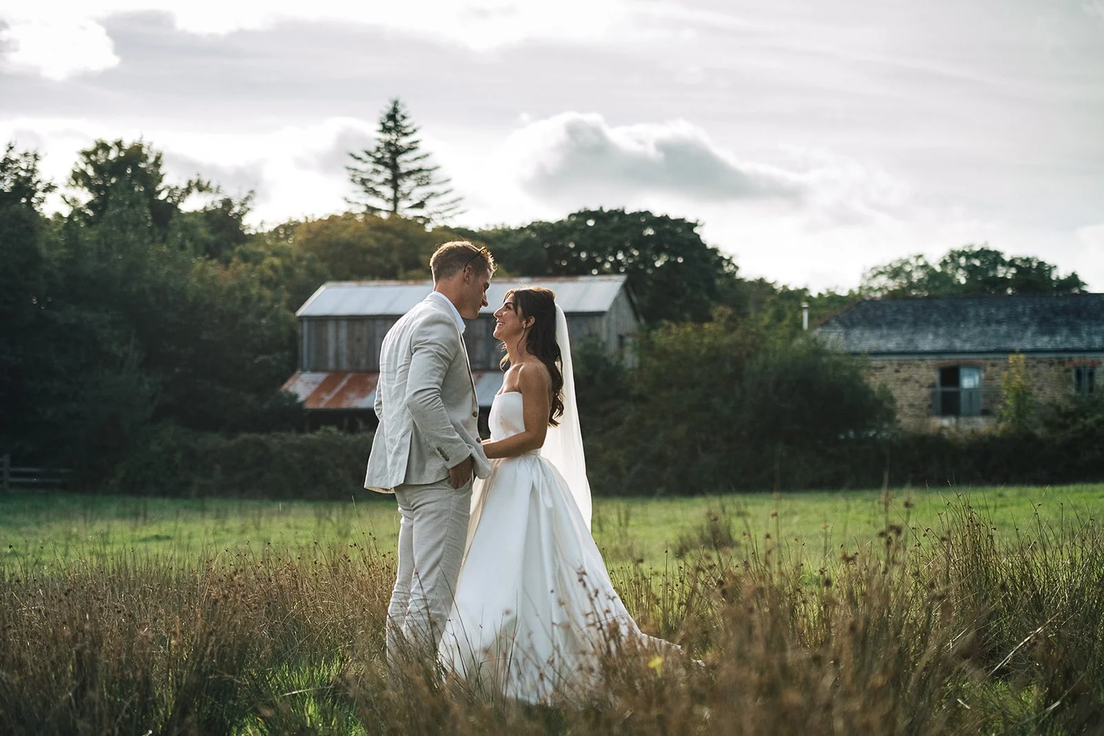 Bride and groom standing close in a field, facing each other, with trees and old barn structures in the background during daytime.