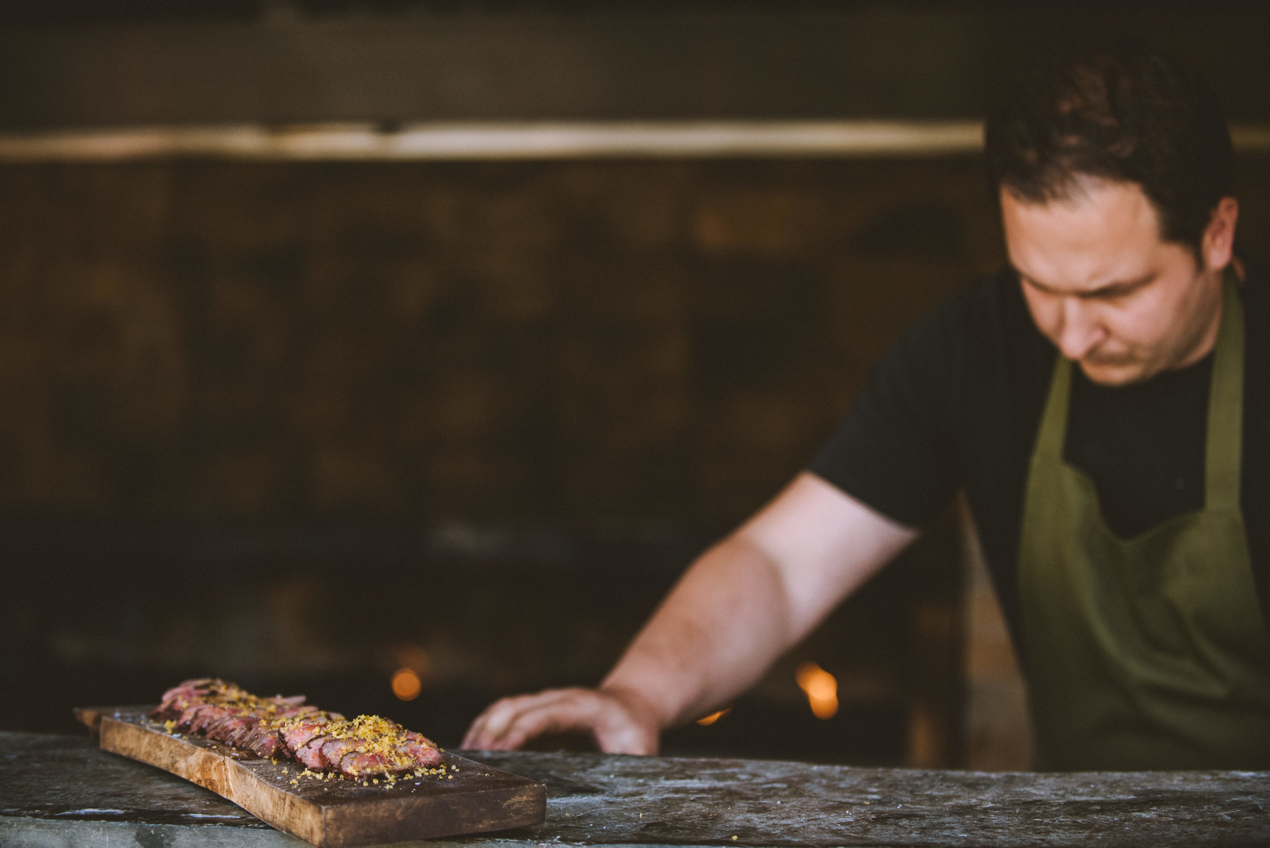 A man with dark hair prepares a dish of sliced meat on a wooden board in a dimly lit kitchen.