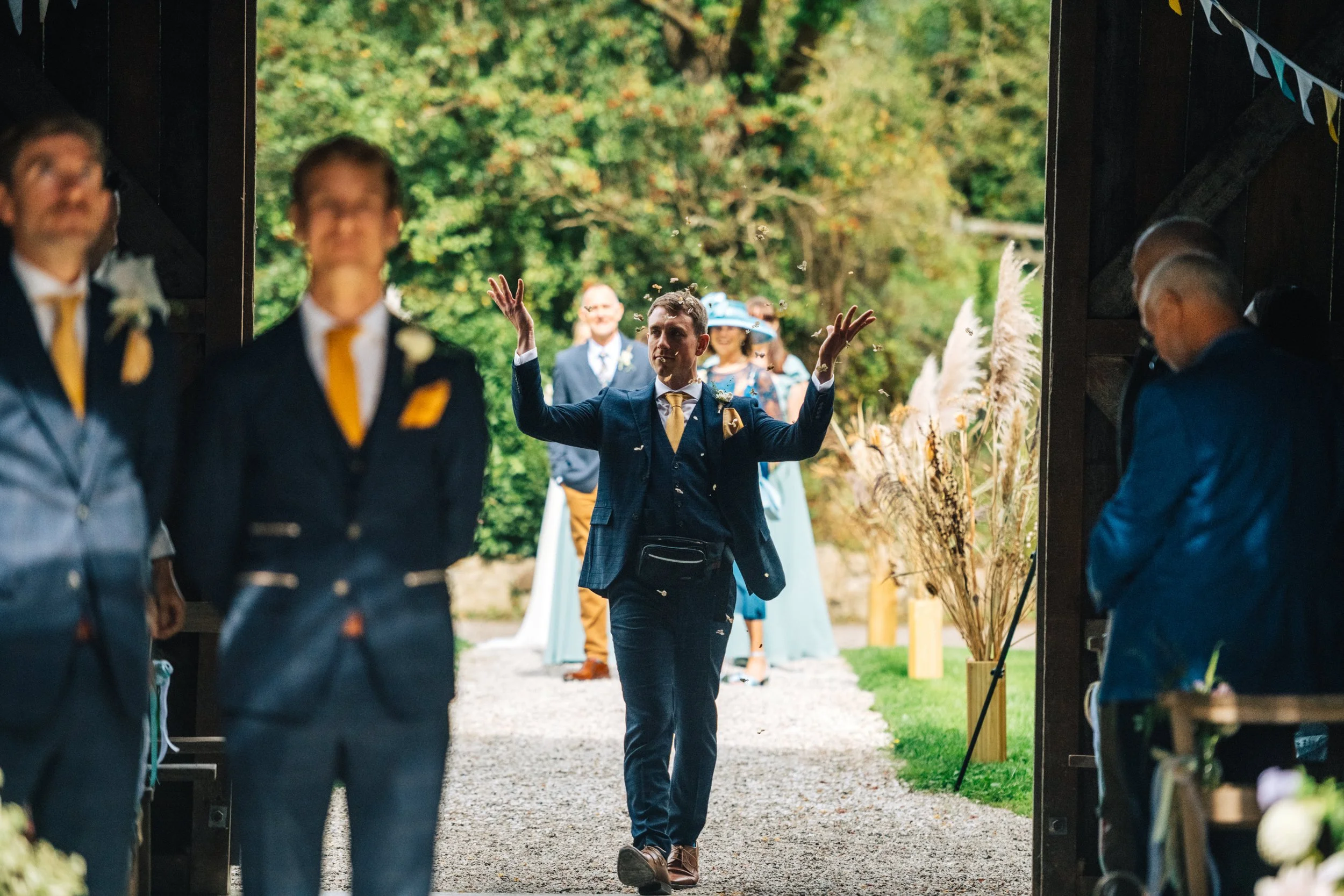 Groom in navy suit walking down aisle with arms raised, wedding guests in background, at outdoor wedding ceremony.