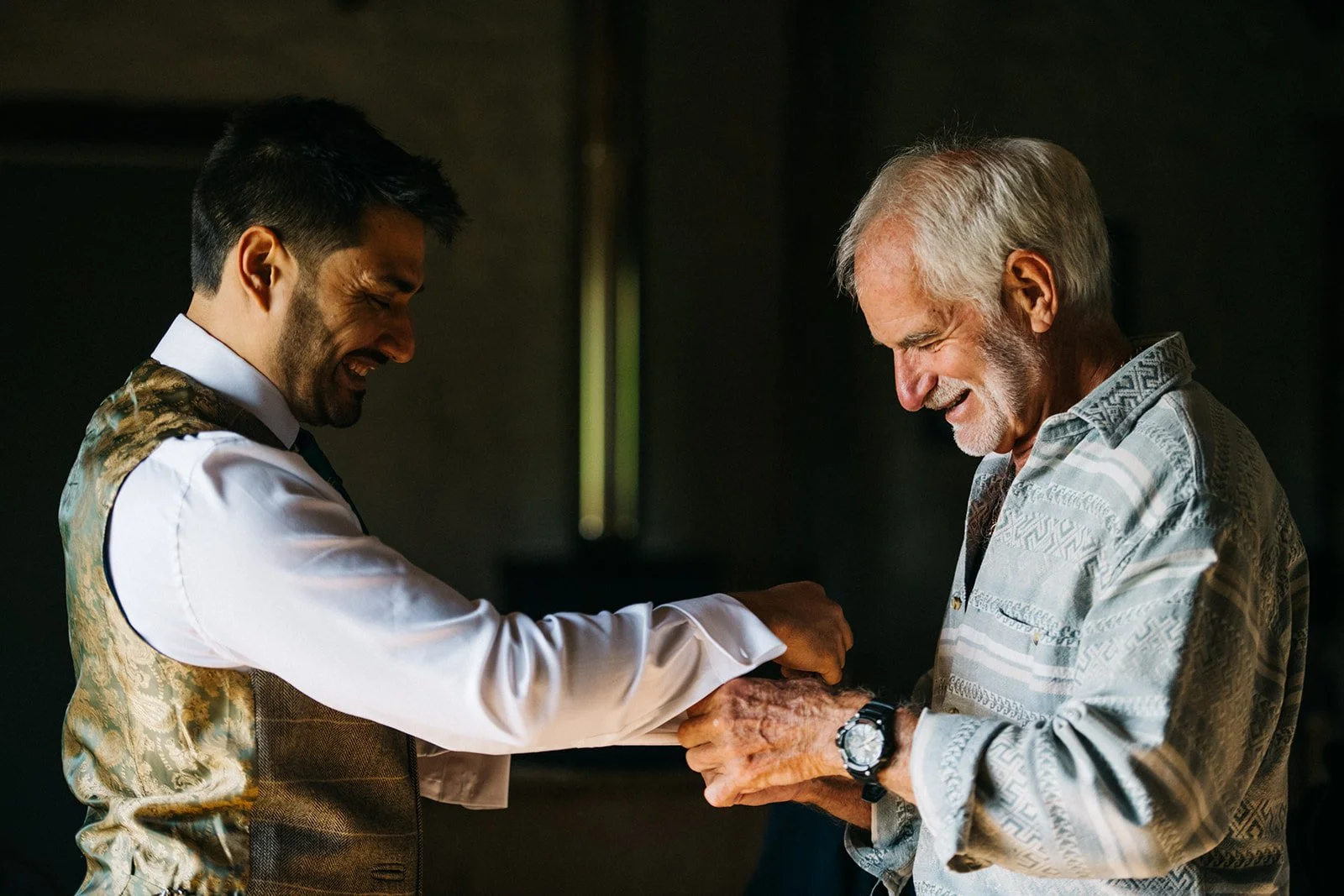 A smiling young man in a vest and white shirt interacts with an older man in a patterned shirt, sharing a joyful moment.