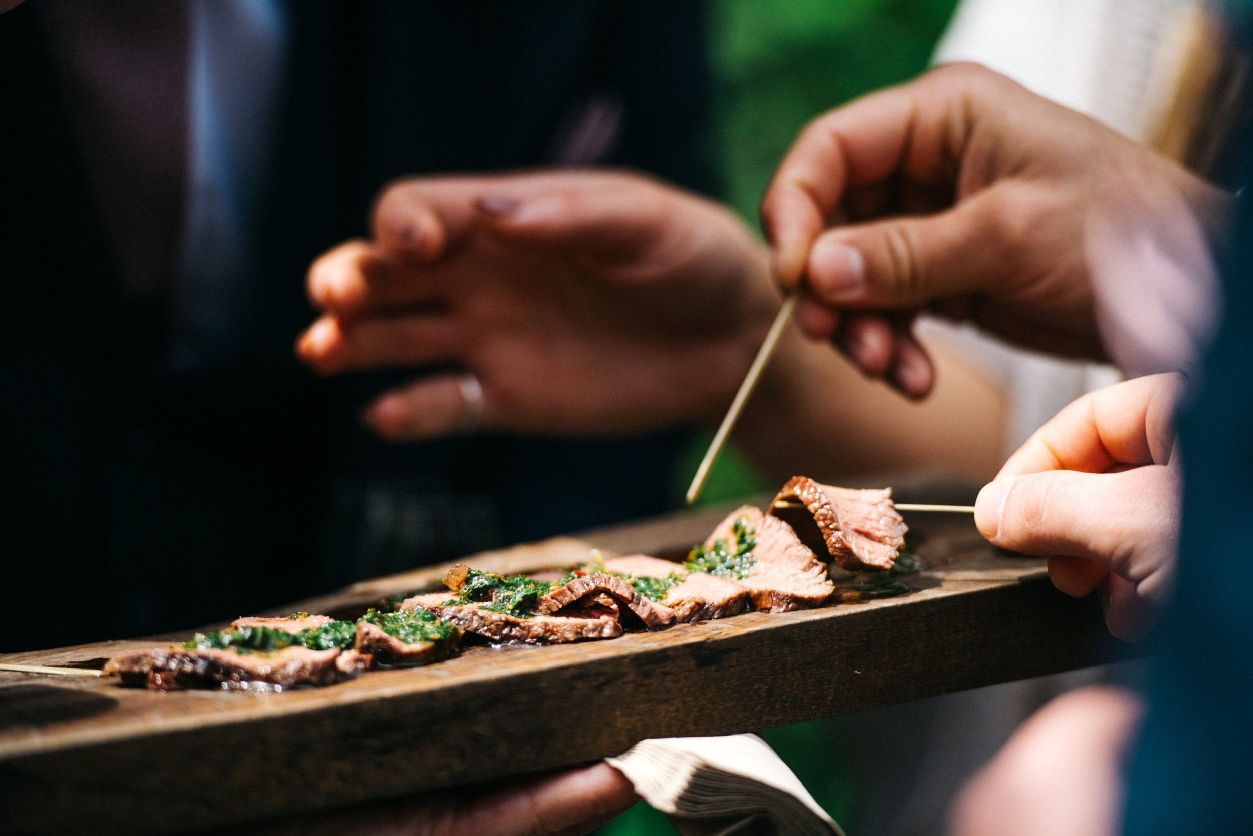 Hands slicing cooked steak garnished with herbs on a wooden serving platter.