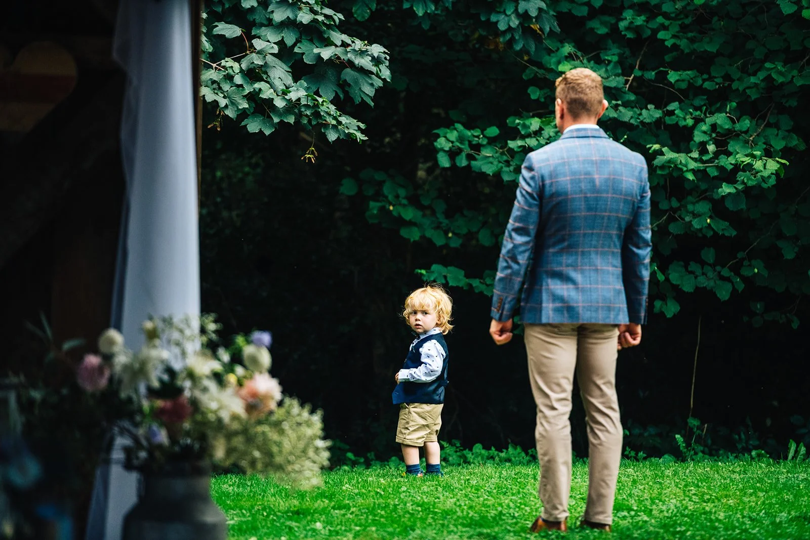 A man in a blue checkered blazer and beige pants stands on a green grassy lawn, looking at a young boy with curly blond hair, who is wearing a navy vest, white shirt with blue patterns, tan shorts, and blue shoes. They are in front of a background of