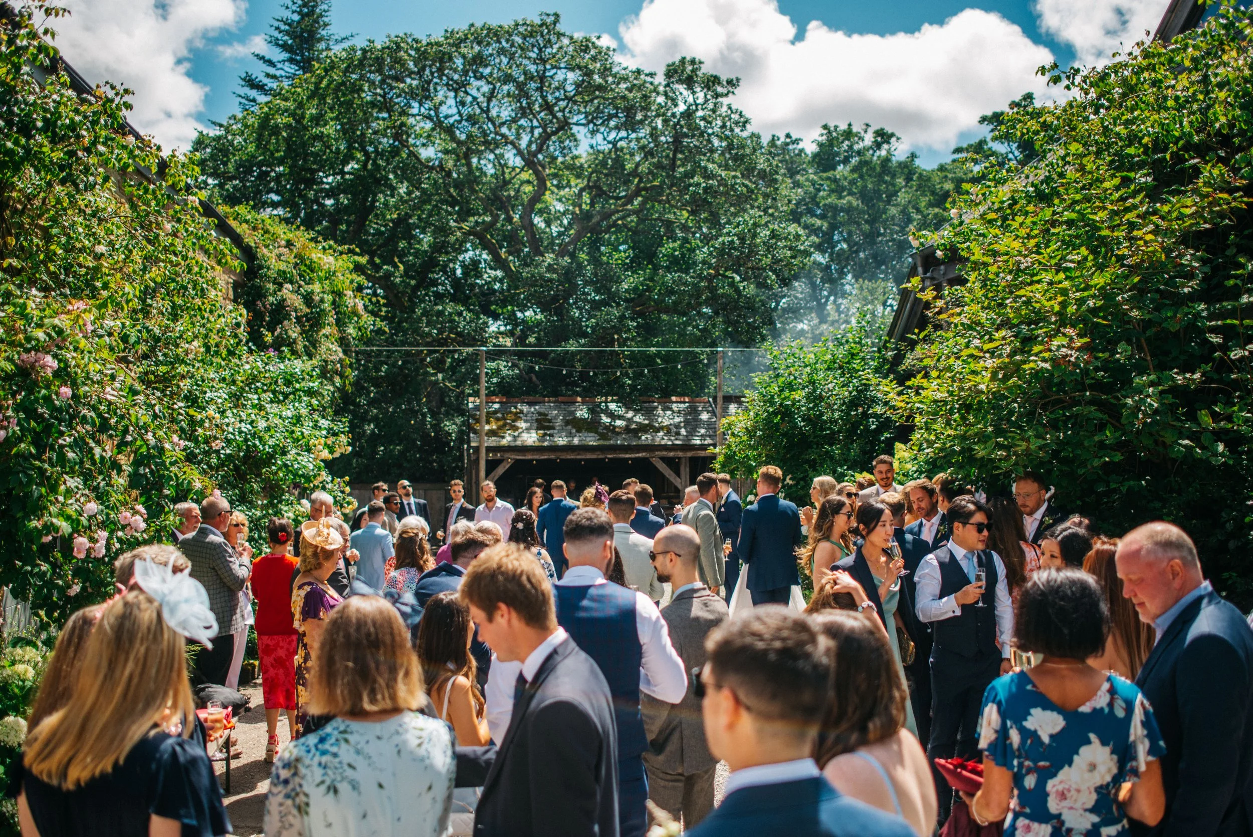 Group of people socializing outdoors in a garden at a daytime event or party, with lush trees and bushes surrounding them.