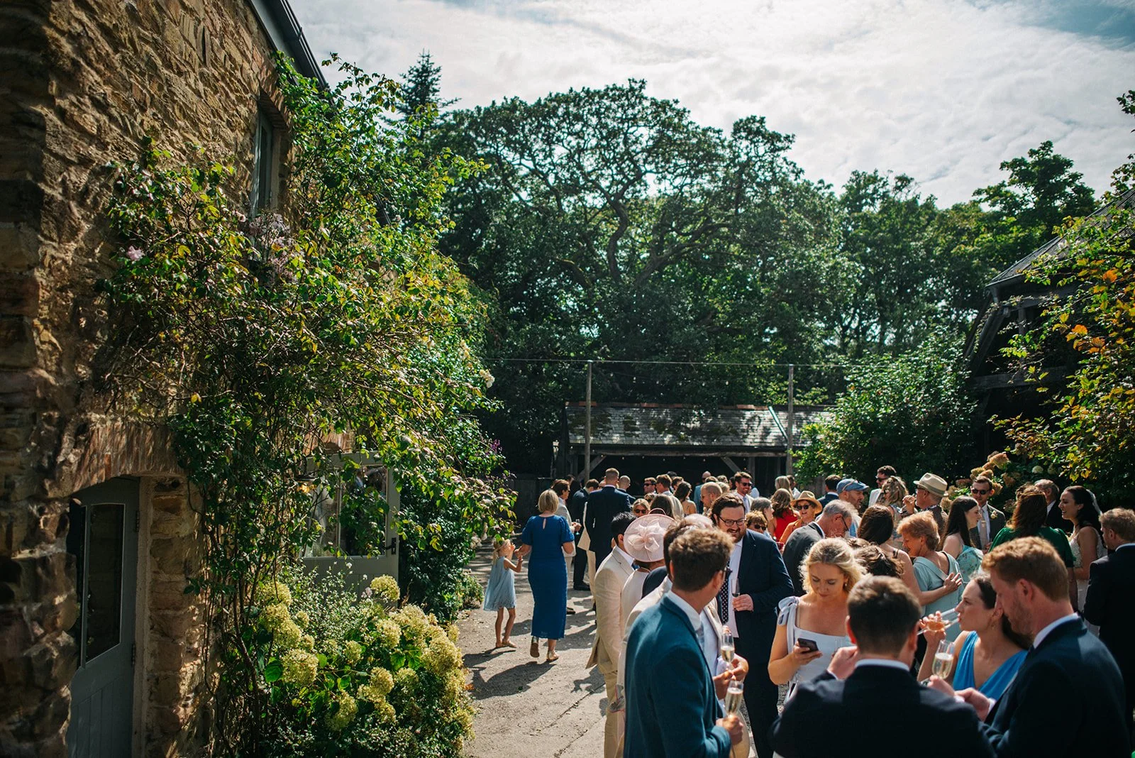 A large outdoor gathering of people in formal attire at a garden party or wedding reception, with greenery, trees, and old buildings in the background on a partly cloudy day.