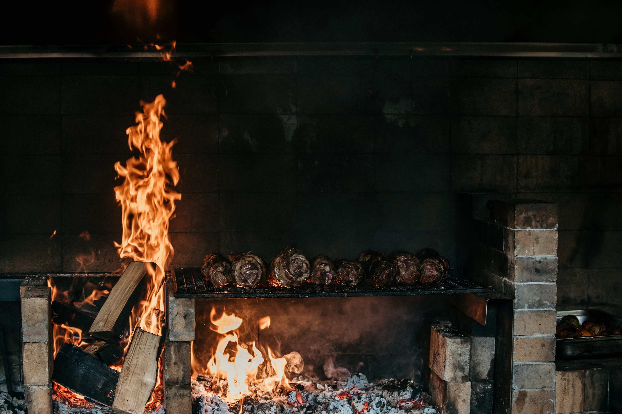 Meat being grilled over an open flame in a brick and metal barbecue grill.
