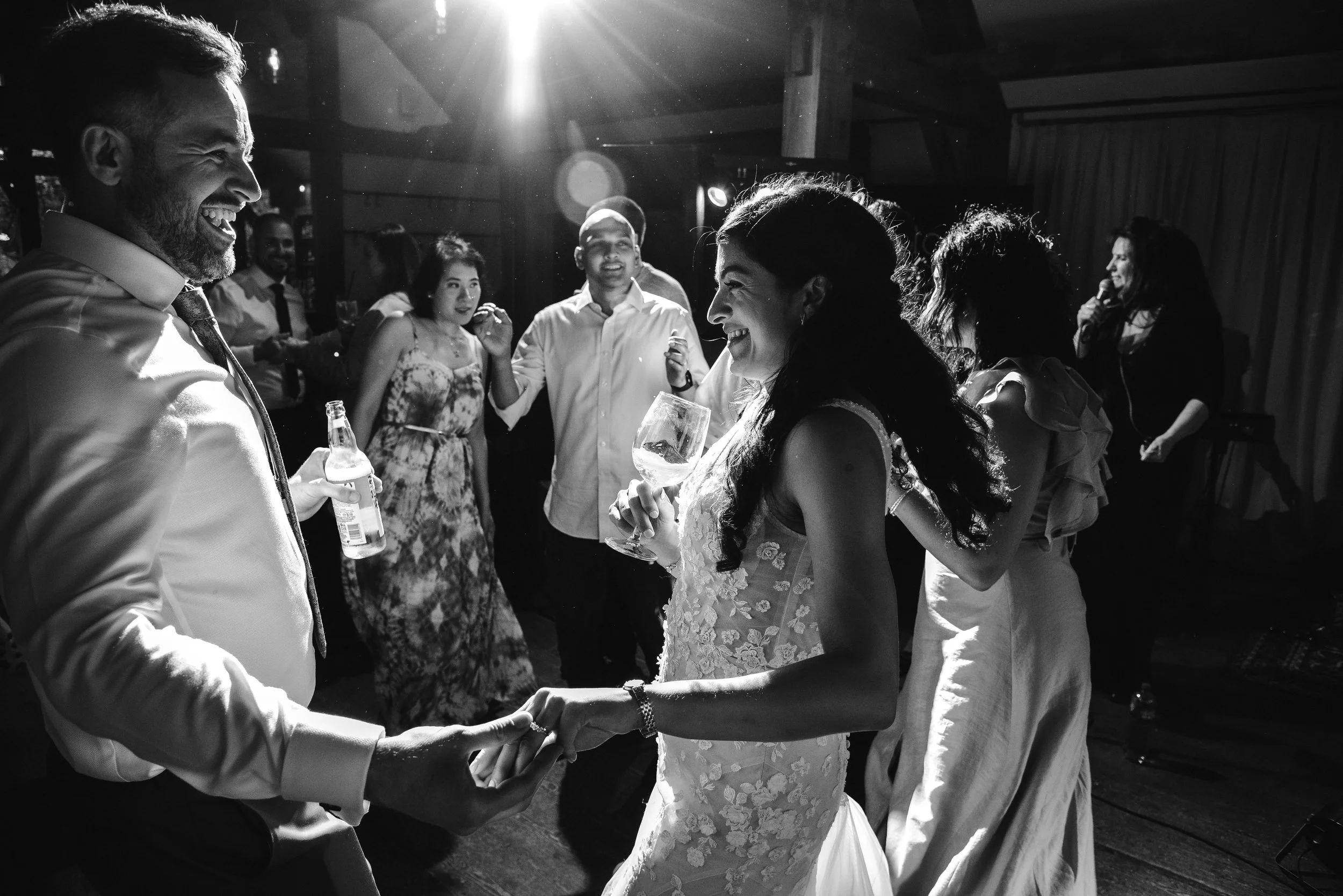 Black and white photo of a wedding reception with a bride and groom dancing and holding hands, surrounded by smiling guests, some holding drinks, in a dimly lit venue.
