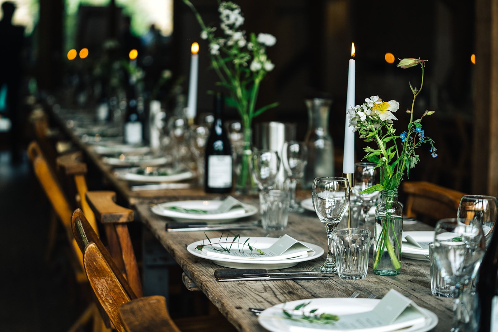 A rustic wooden dining table set for a formal dinner with white plates, glassware, and cutlery, decorated with vases of flowers and lit candles.