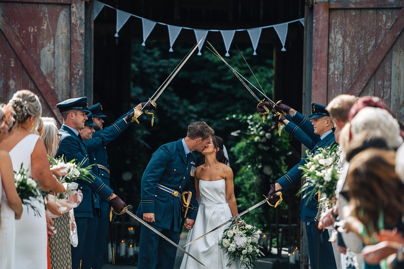 Couple kissing under ceremony arch with military honor guards in uniform, holding swords, at outdoor wedding.