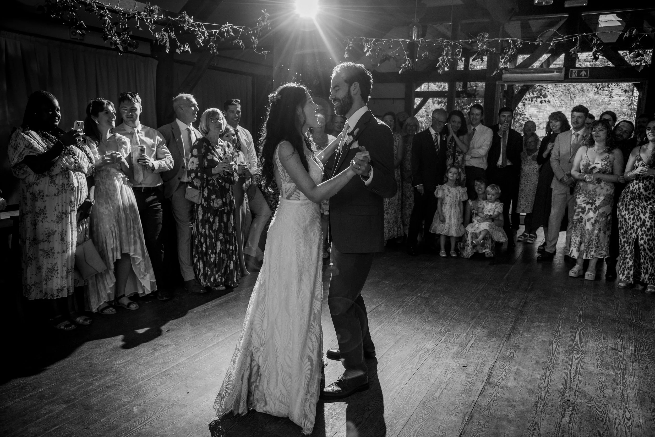 A black and white photo of a bride and groom dancing at their wedding reception, surrounded by guests watching.