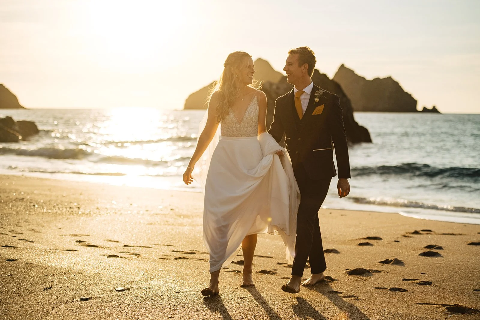 A bride and groom walking barefoot on a beach at sunset, looking at each other and smiling.