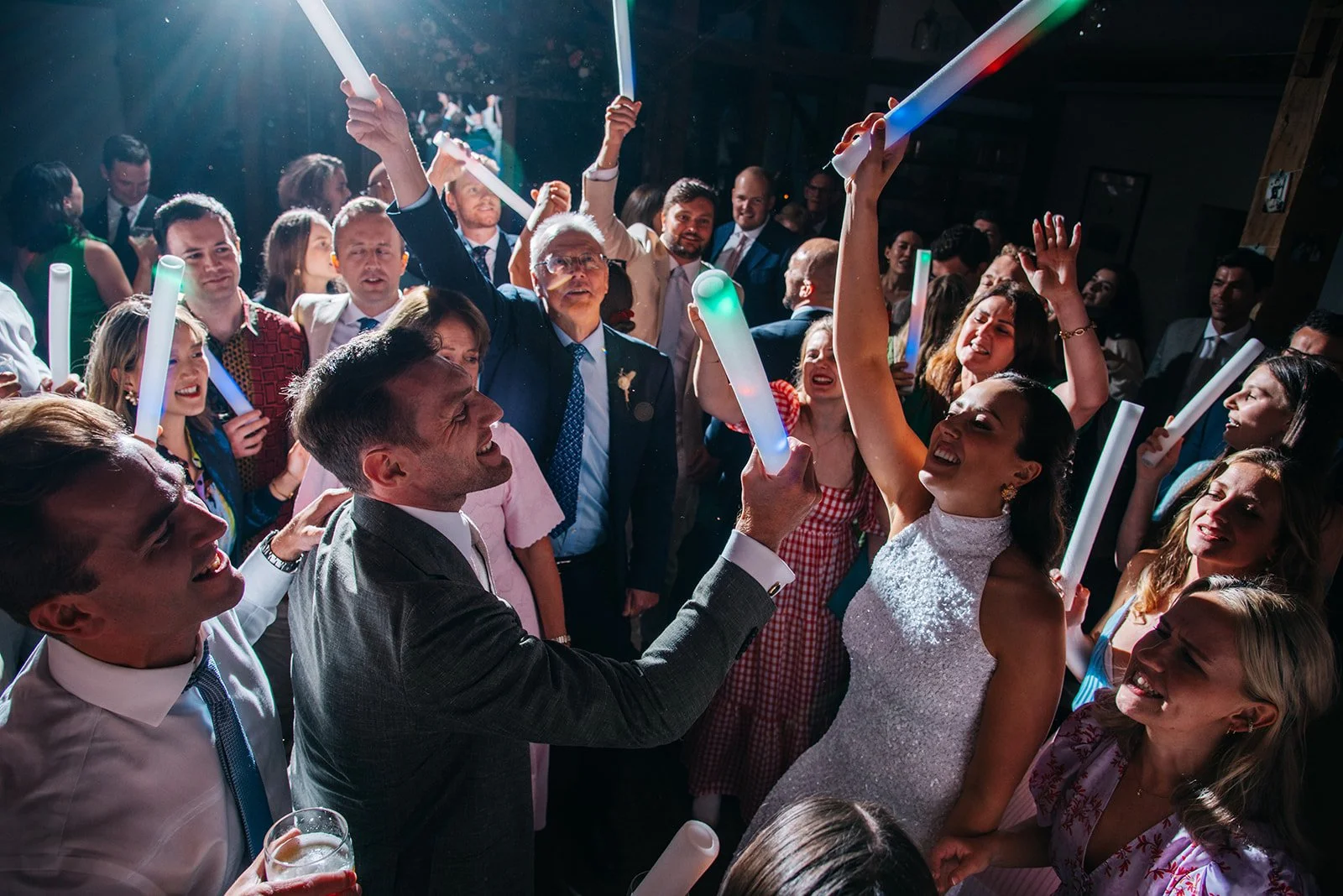 A group of people at a celebration, holding illuminated glow sticks and dancing together in a lively indoor setting.