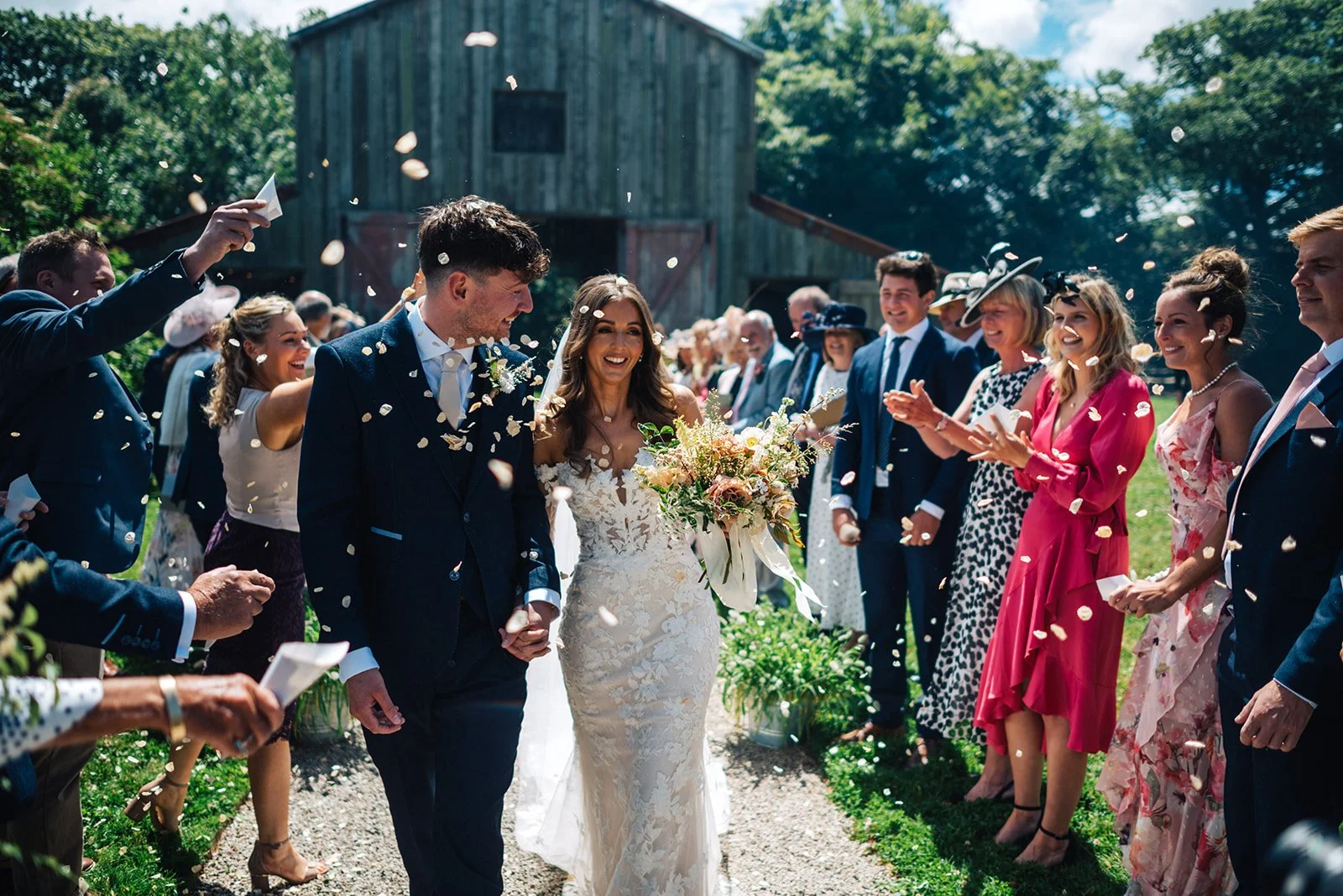 A bride and groom walking down the aisle while friends and family celebrate and throw flower petals in a sunny outdoor wedding ceremony.