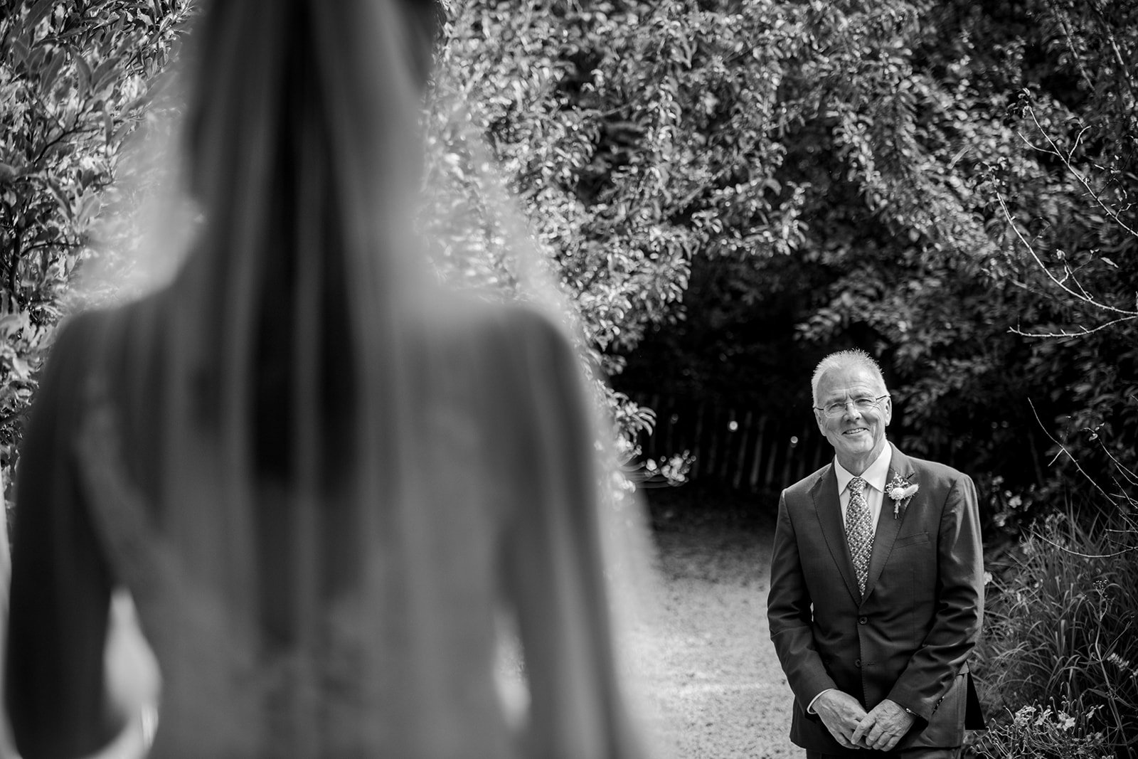 An older man in a suit standing outdoors, smiling, with trees and a fence in the background. A blurred figure of a woman in the foreground, facing him.