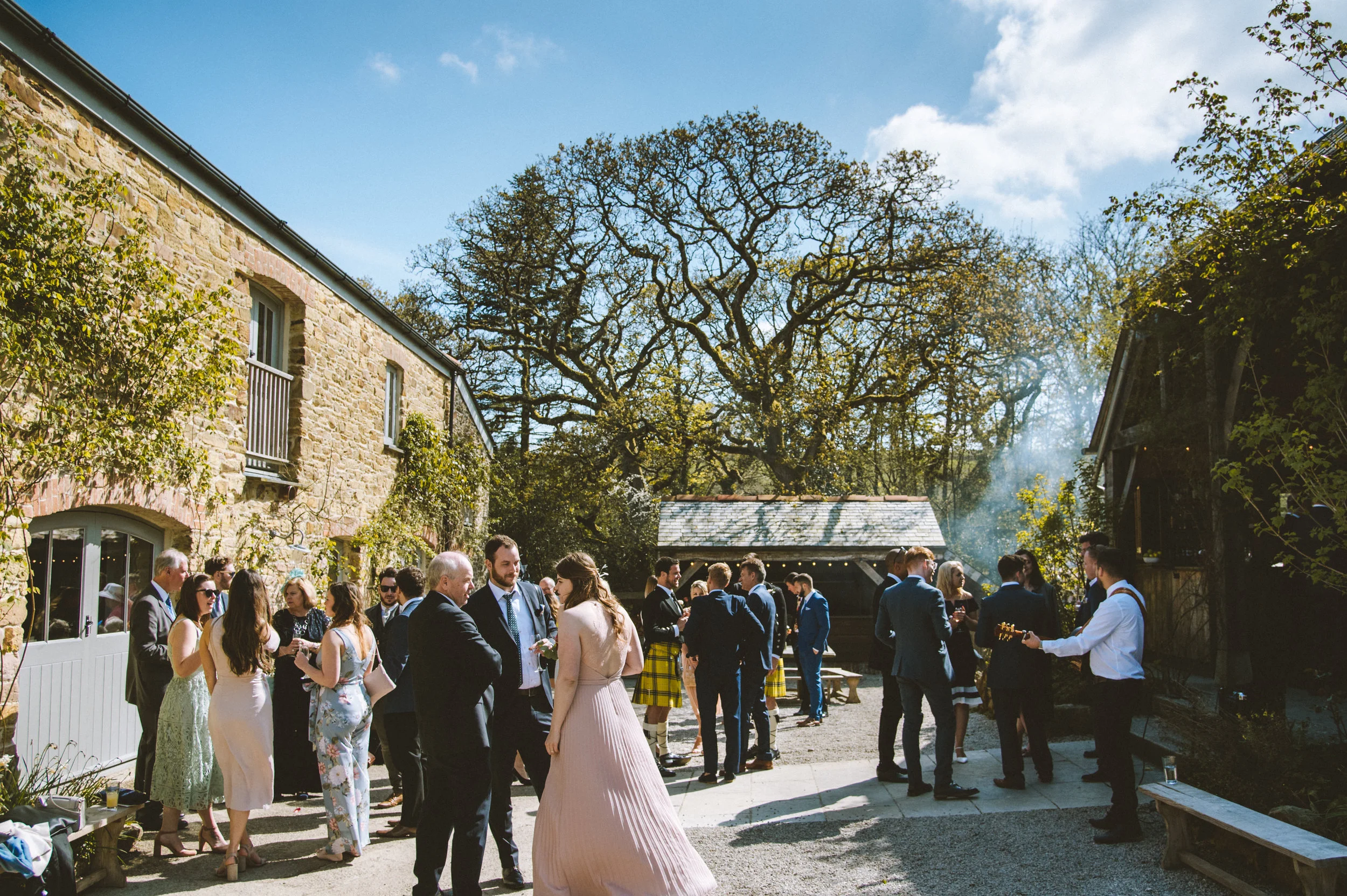 Guests in formal attire socializing outdoors at a wedding or party in a courtyard with stone buildings, trees, and blue sky.