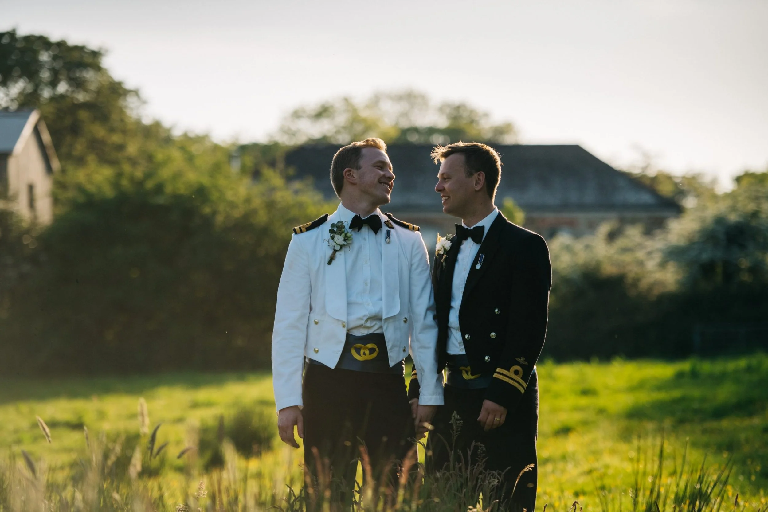Two men in military dress uniforms holding hands and smiling at each other in a field with green grass and trees, during sunset.