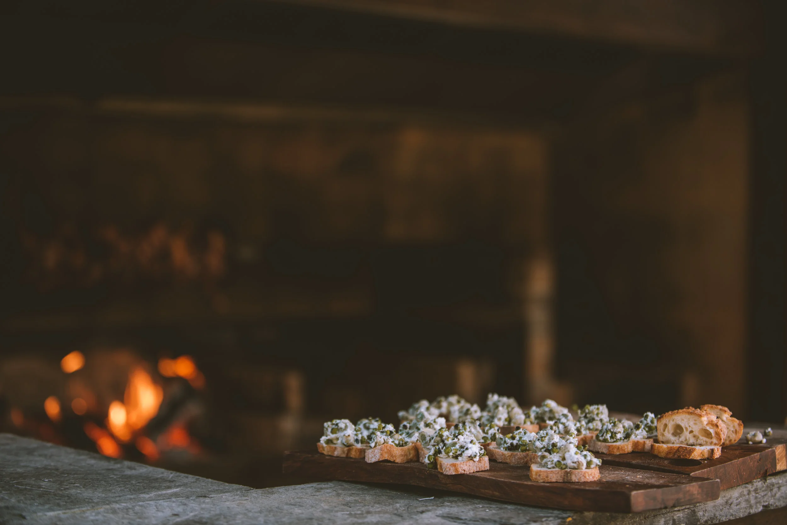 Appetizer crostini topped with white cheese and herbs on a wooden cutting board in front of a fireplace.