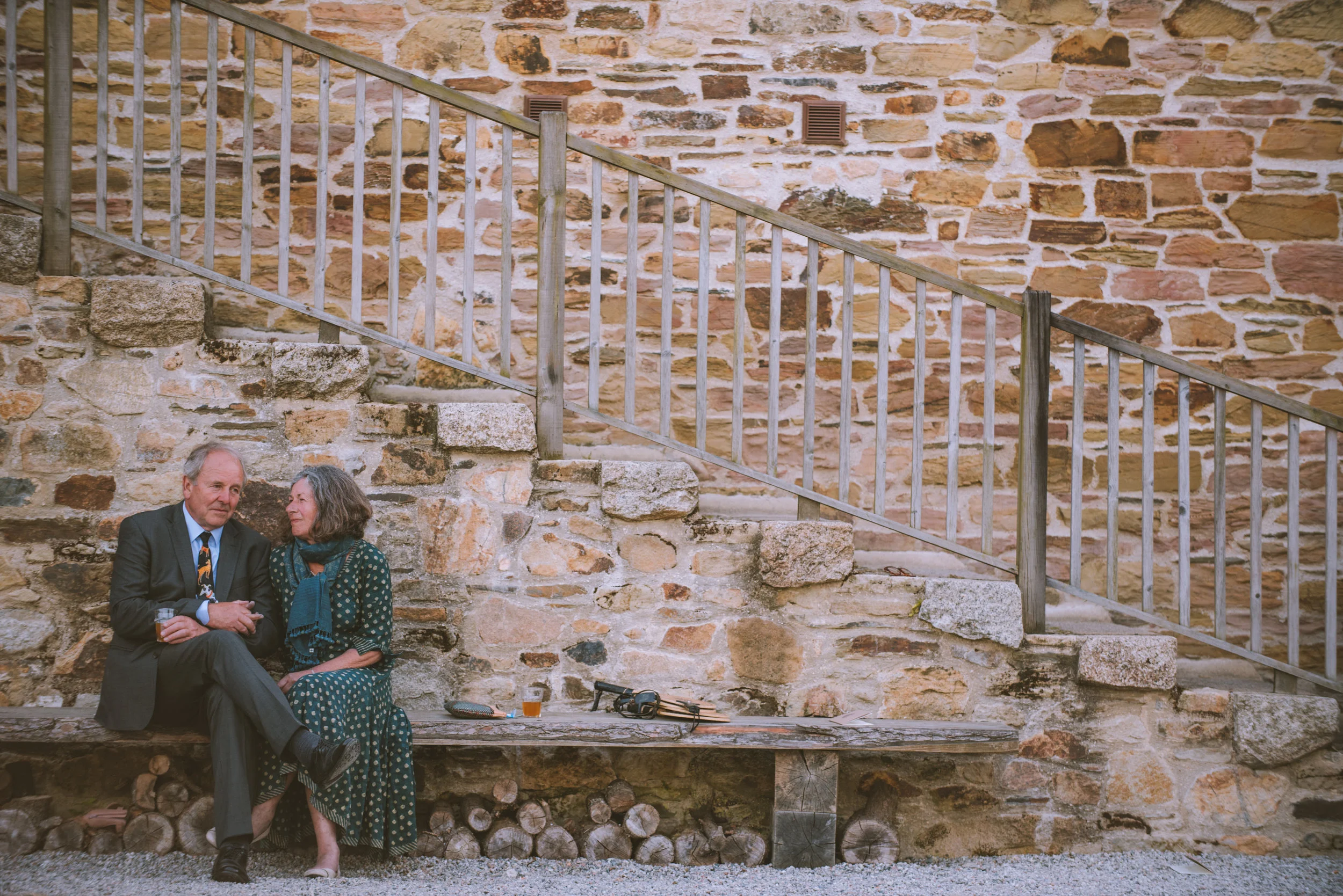 An elderly man and woman sitting on a wooden bench against a stone wall, engaging in conversation with drinks and personal items nearby.
