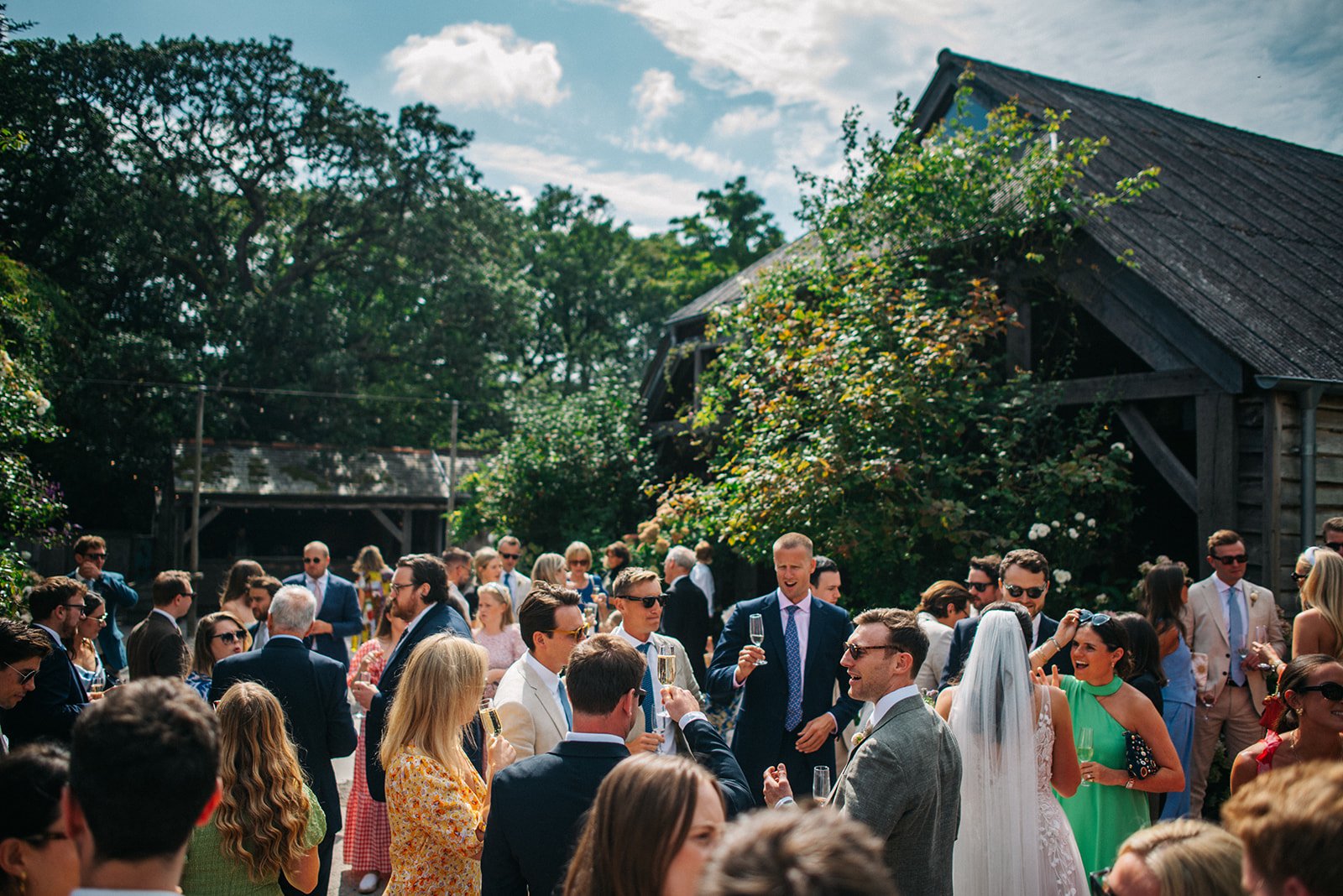 Guests celebrating outdoors at a wedding reception, some holding glasses of champagne, on a sunny day with a blue sky, trees, and rustic buildings in the background.