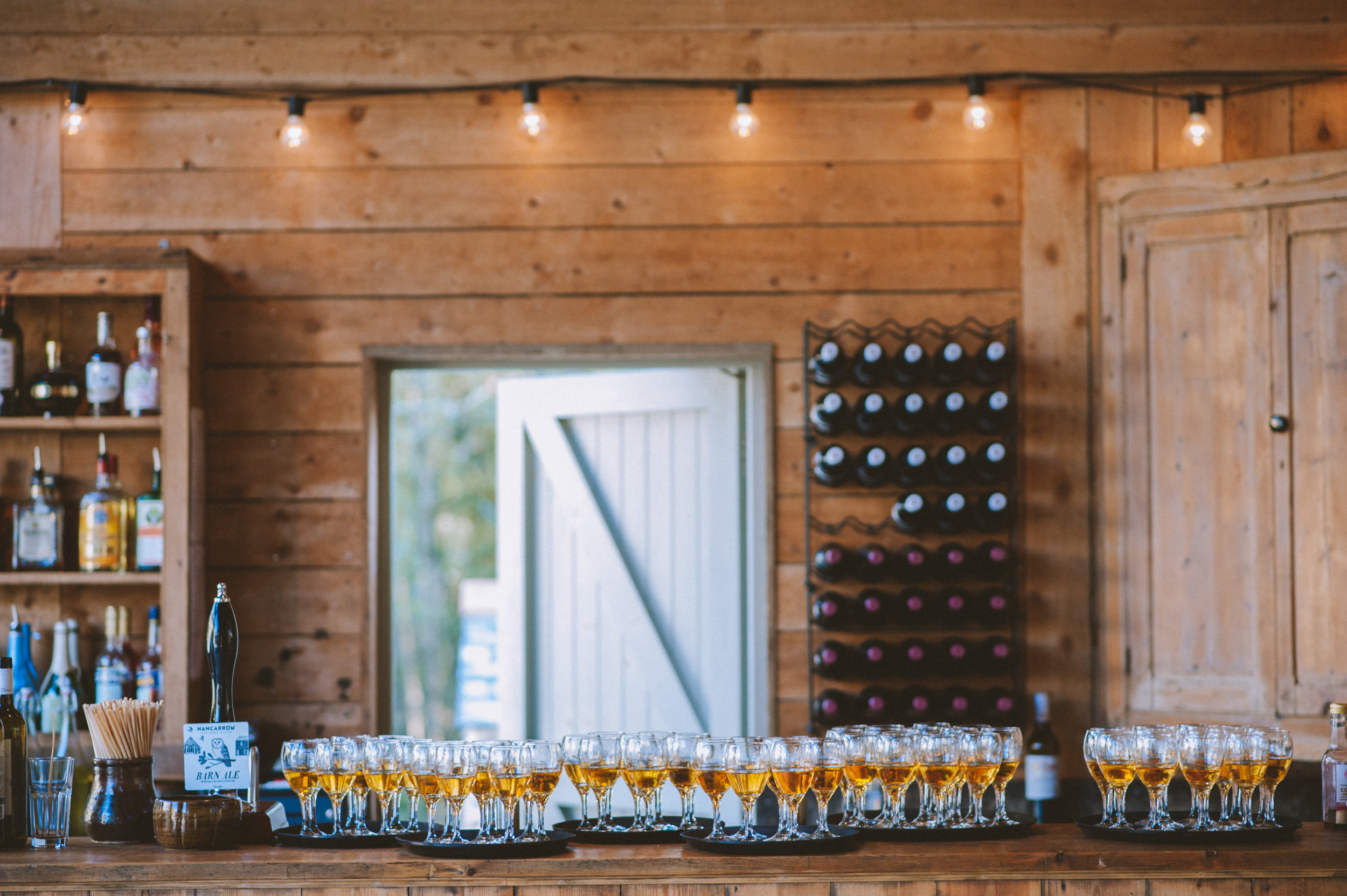 A rustic wooden bar with multiple glasses of beer, bottles of alcohol, and a bottle opener, with a window and shelves of wine bottles in the background.