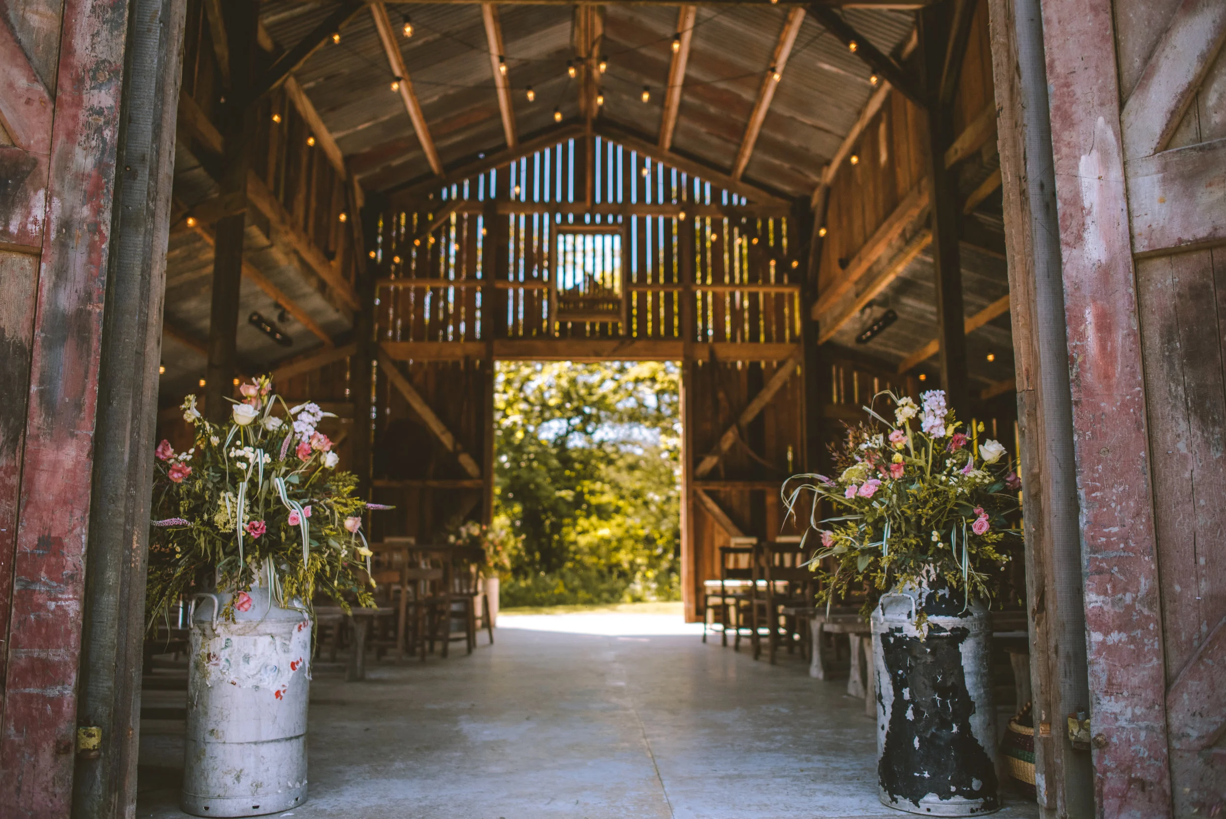 Interior of a rustic barn with wooden walls and ceiling, decorated with string lights, featuring two large vases with pink and white flowers at the entrance, and chairs arranged inside for an event.